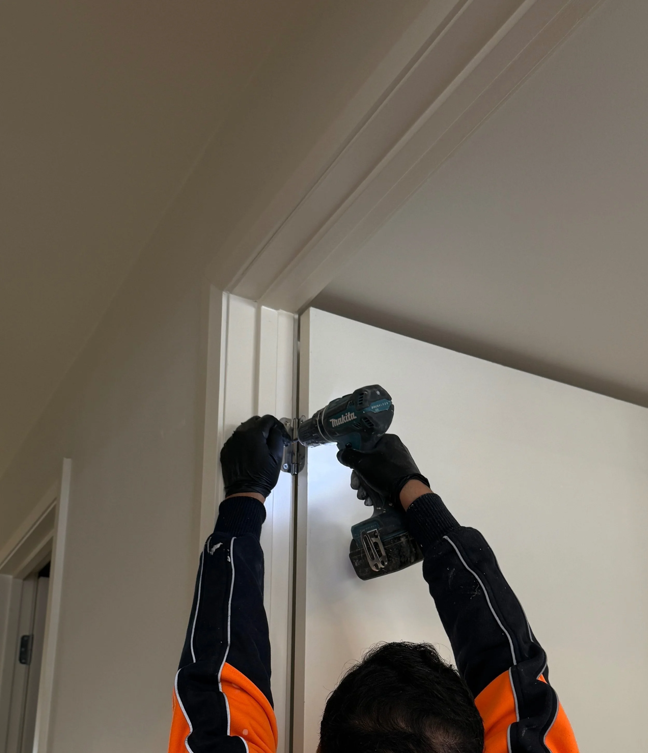 CBG construction pty ltd person using a cordless drill to install or repair a door frame in an interior space, wearing black gloves and an orange and black jacket.