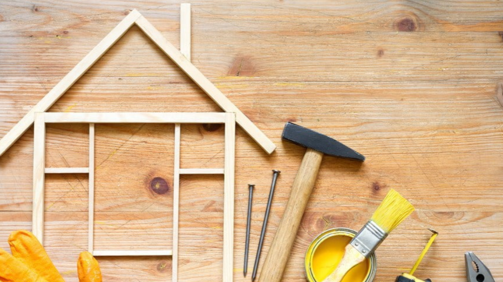 CBG construction solution Woodworking tools and materials arranged on a wooden surface, including a small house frame, hammer, nails, paintbrush, yellow paint, gloves, and a level.