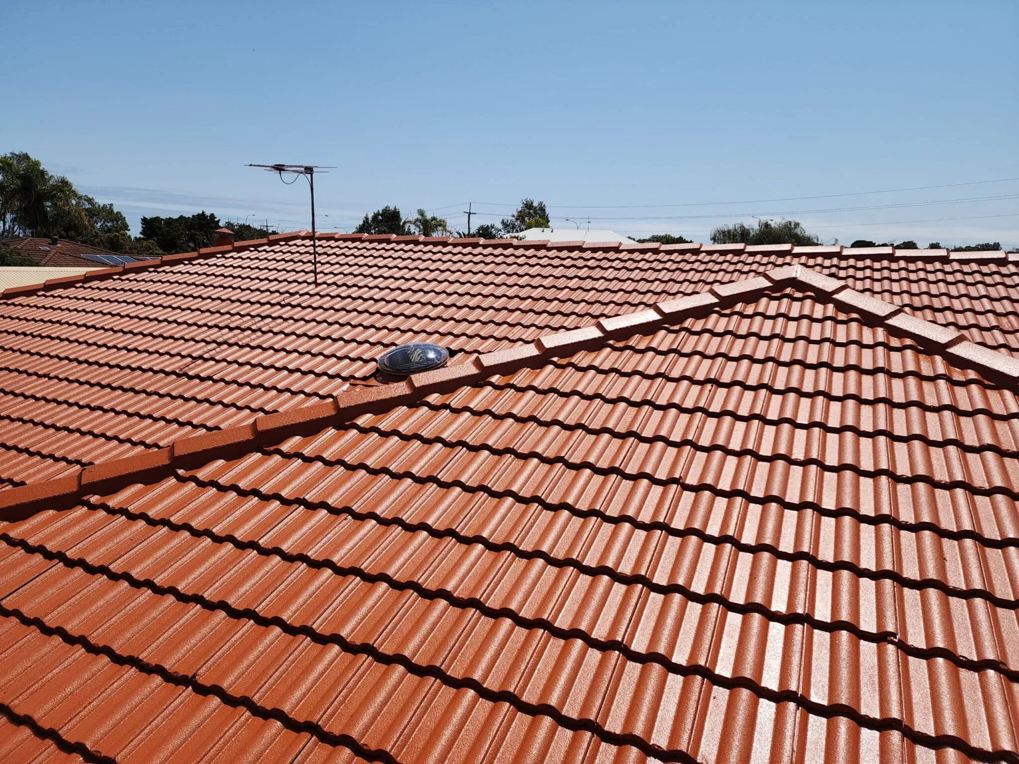 CBG work Close-up view of a red tiled roof with a solar light fixture and an antenna on top, under a clear blue sky, with trees and other rooftops in the background.