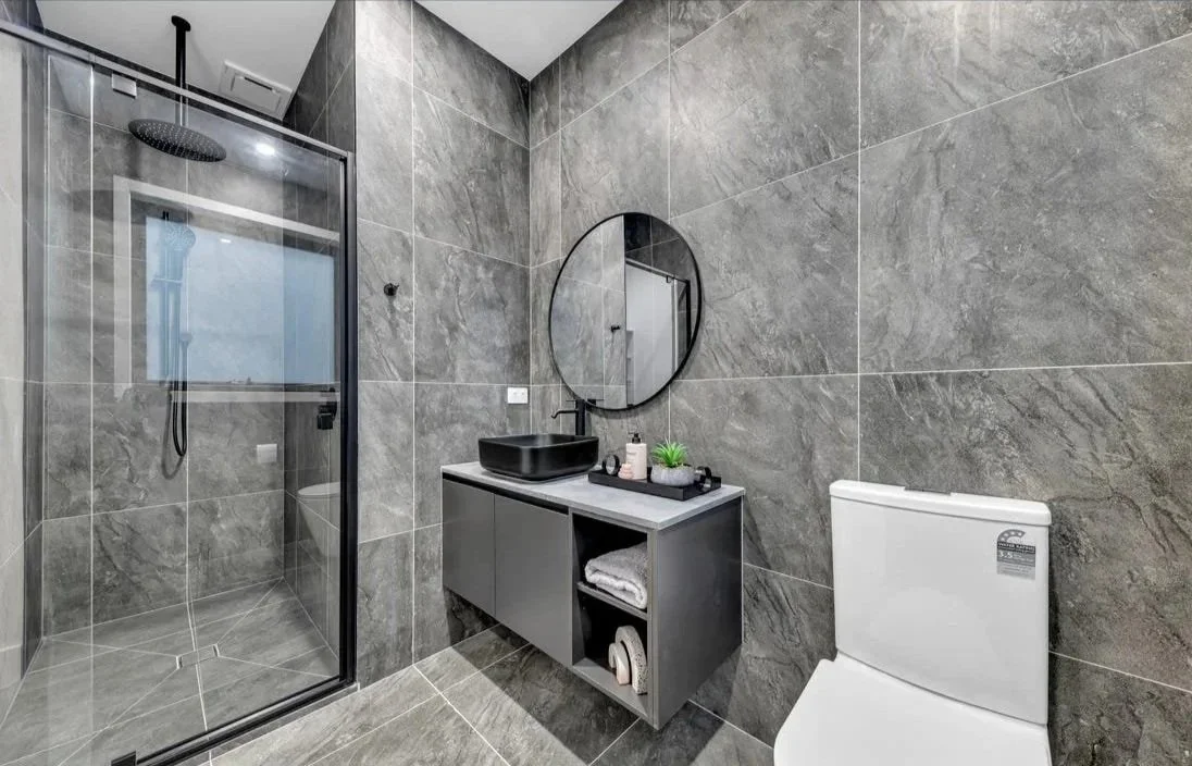 CBG CONSTRUCTION SOLUTIONS PTY LTD BEST COMPANY IN MELBOURNE Modern gray tiled bathroom with walk-in shower, black vessel sink, round mirror, gray vanity with rolled towel and toiletries, and white toilet.