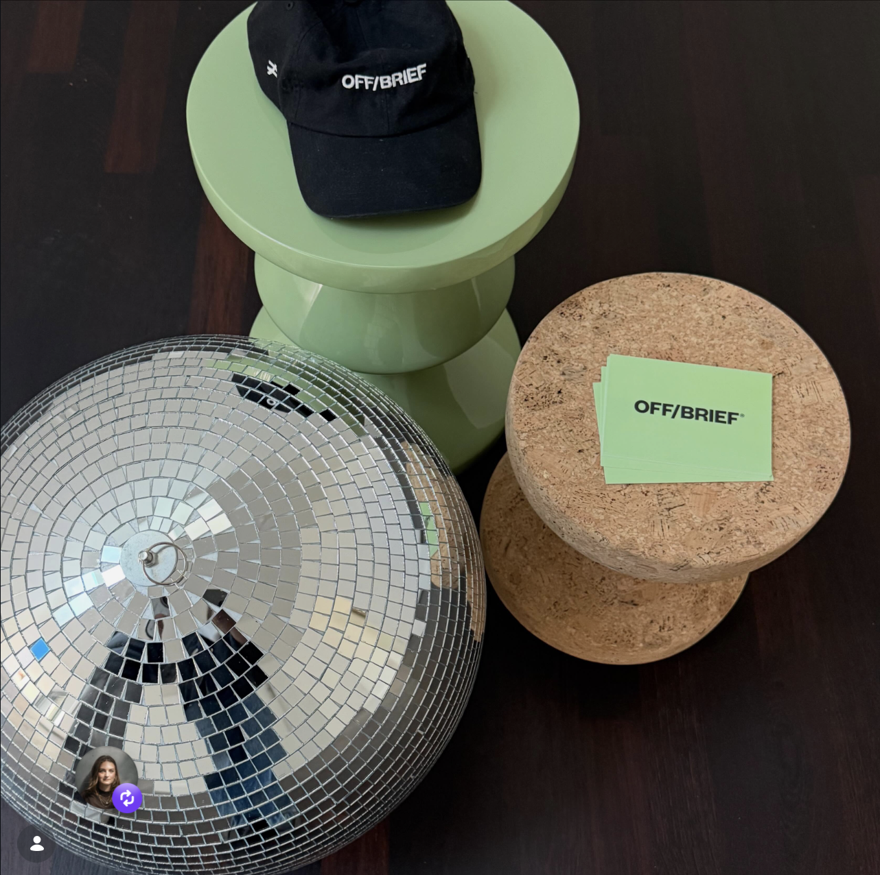 Schwarze Mütze mit weißem Text "OFF/BRIEF" auf einem grünen Hocker, Verpackungen mit dem selben Text auf einem runden Korkhocker, eine große silberne Discokugel auf dem Tisch.