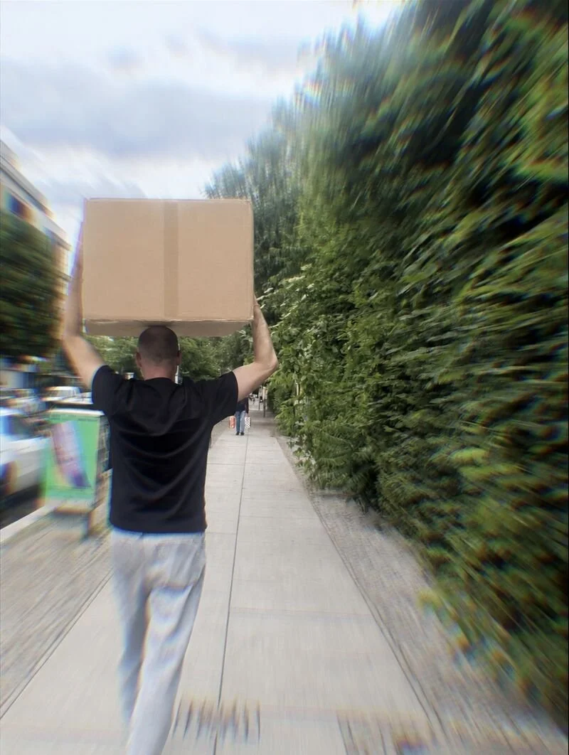 Mann läuft auf Bürgersteig, trägt Karton auf dem Kopf, bunte Bäume neben ihm, verschwommen im Hintergrund.