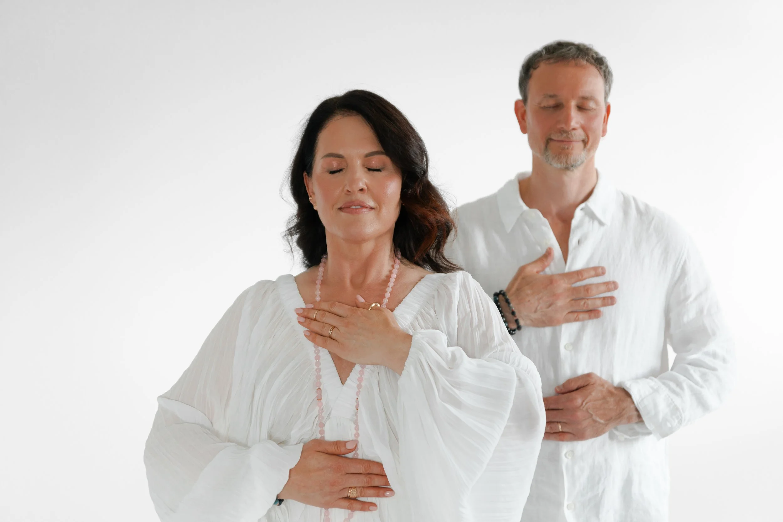 A woman and a man standing with their eyes closed, hands on their chests, in a meditative pose, against a plain white background, wearing white clothing.