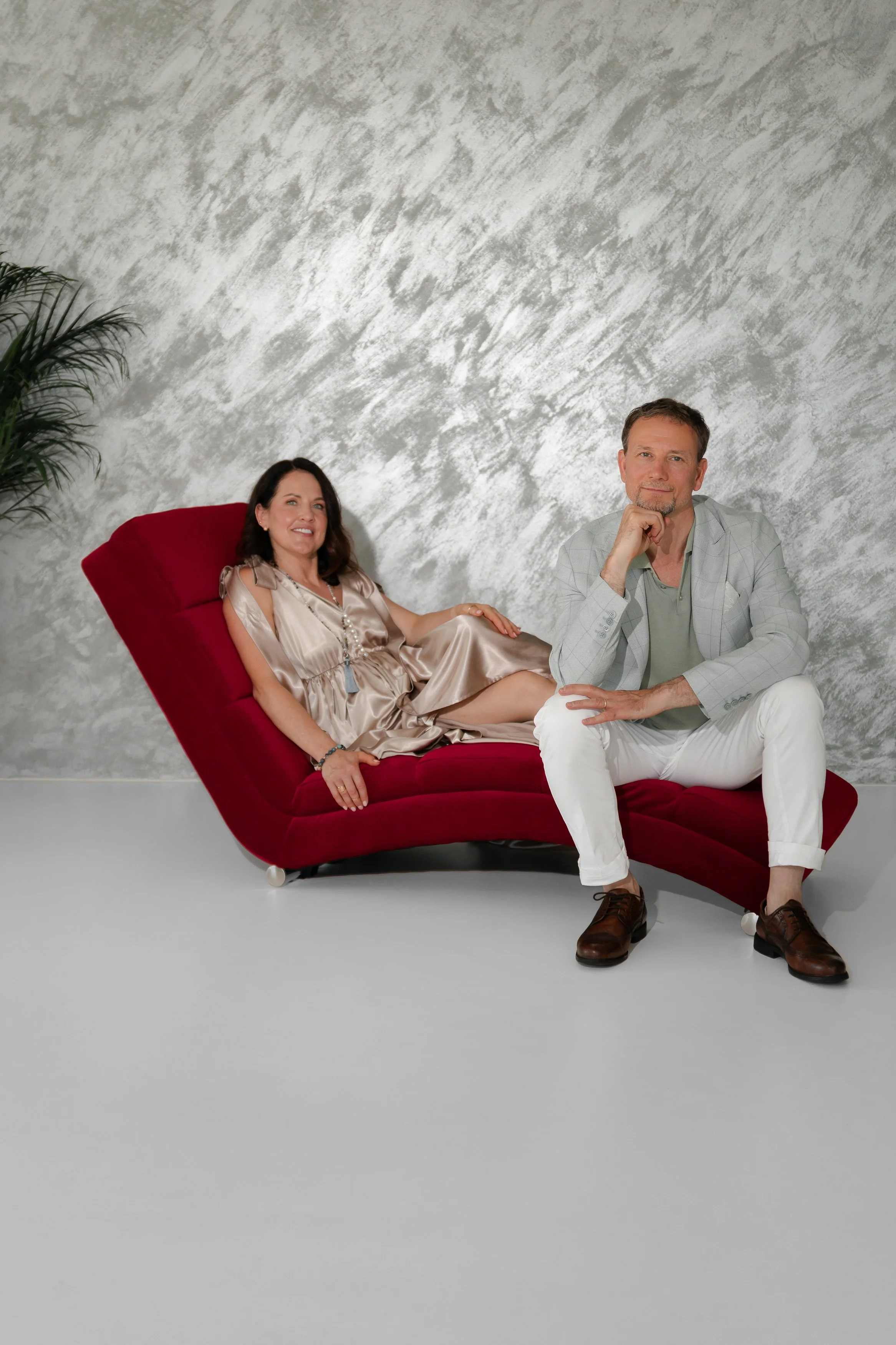 A man and woman posing on a modern red chaise lounge against a mottled gray wall, with a tall green plant in the background.