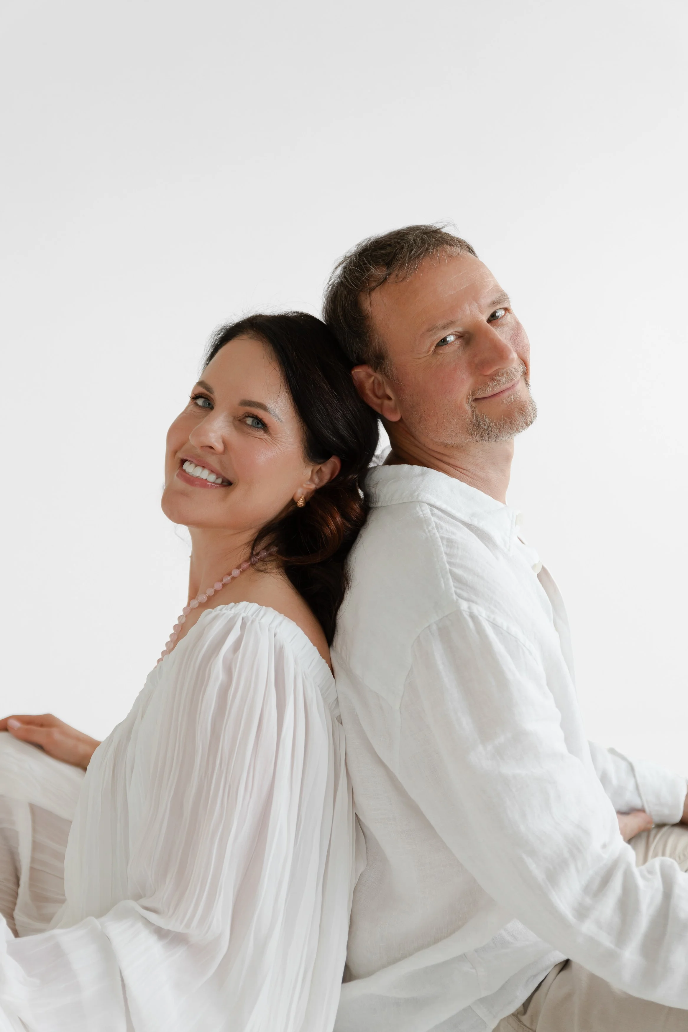 A smiling woman with long dark hair and a man with short light hair and a beard, sitting back-to-back and leaning on each other in a bright, minimalistic setting.