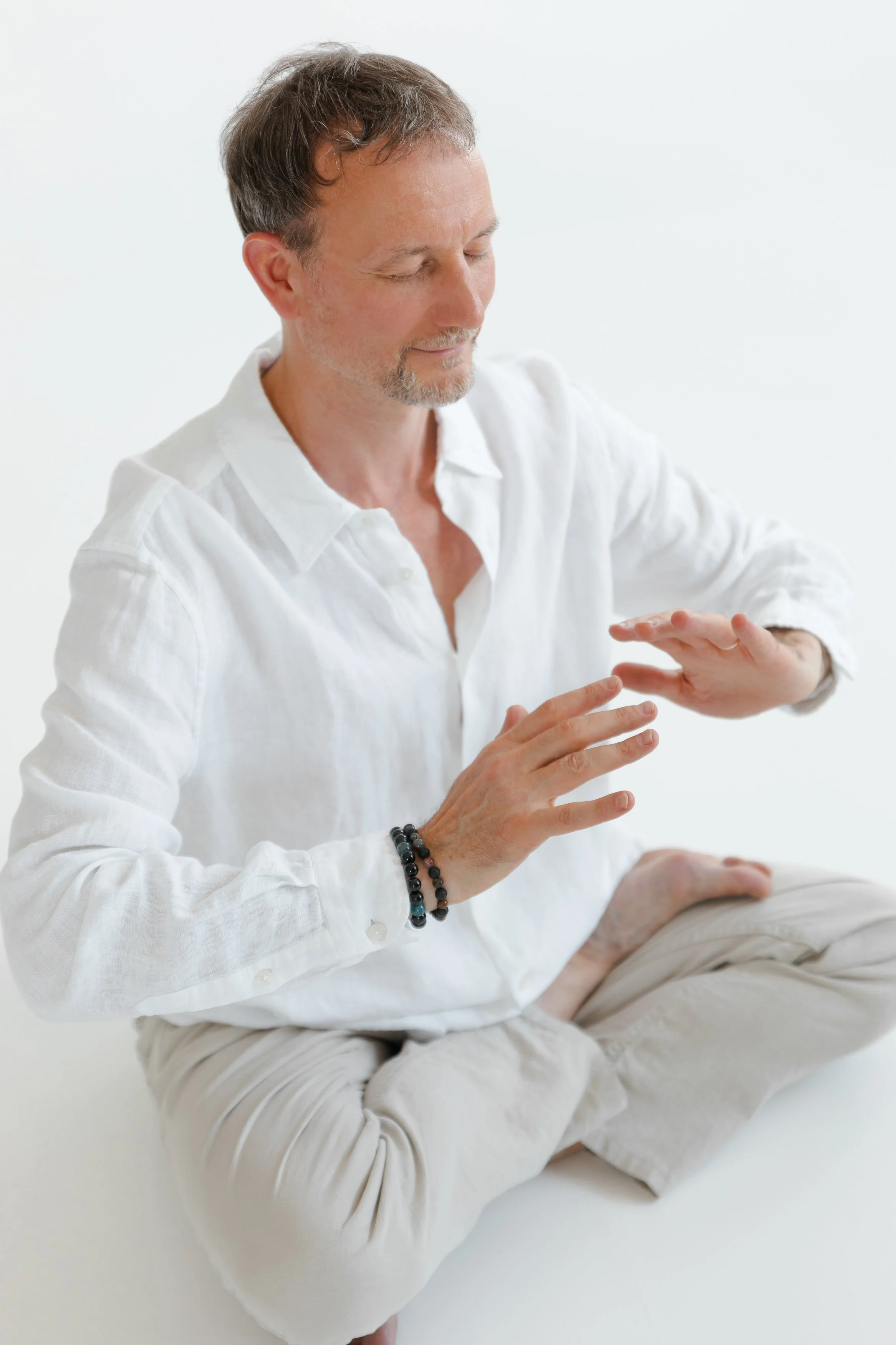 A middle-aged man in a white shirt and beige pants, sitting cross-legged on the floor, with eyes closed and hands raised in a meditative pose against a white background.
