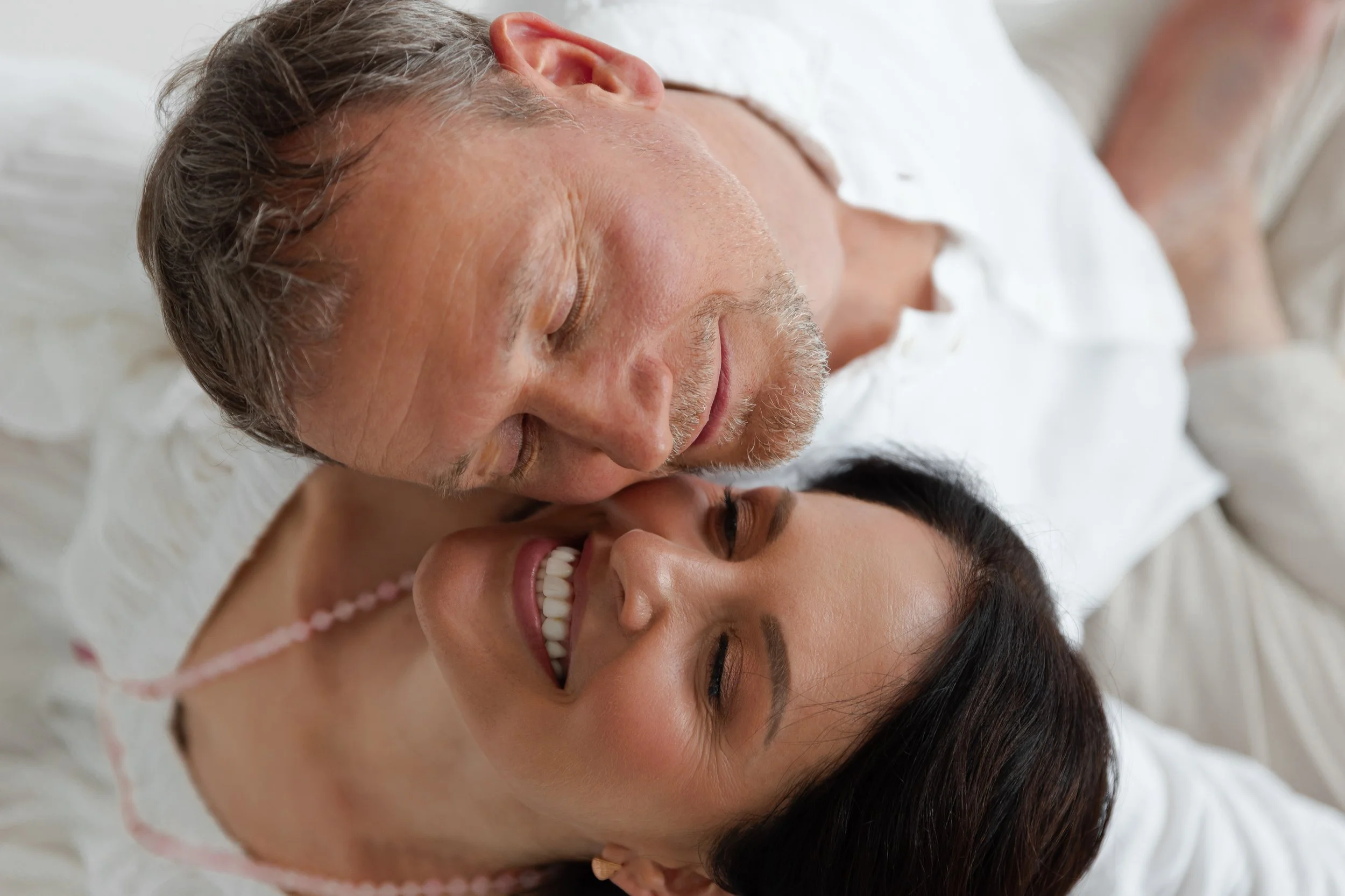 A close-up of an elderly man and a young woman lying on a bed, smiling and touching foreheads.