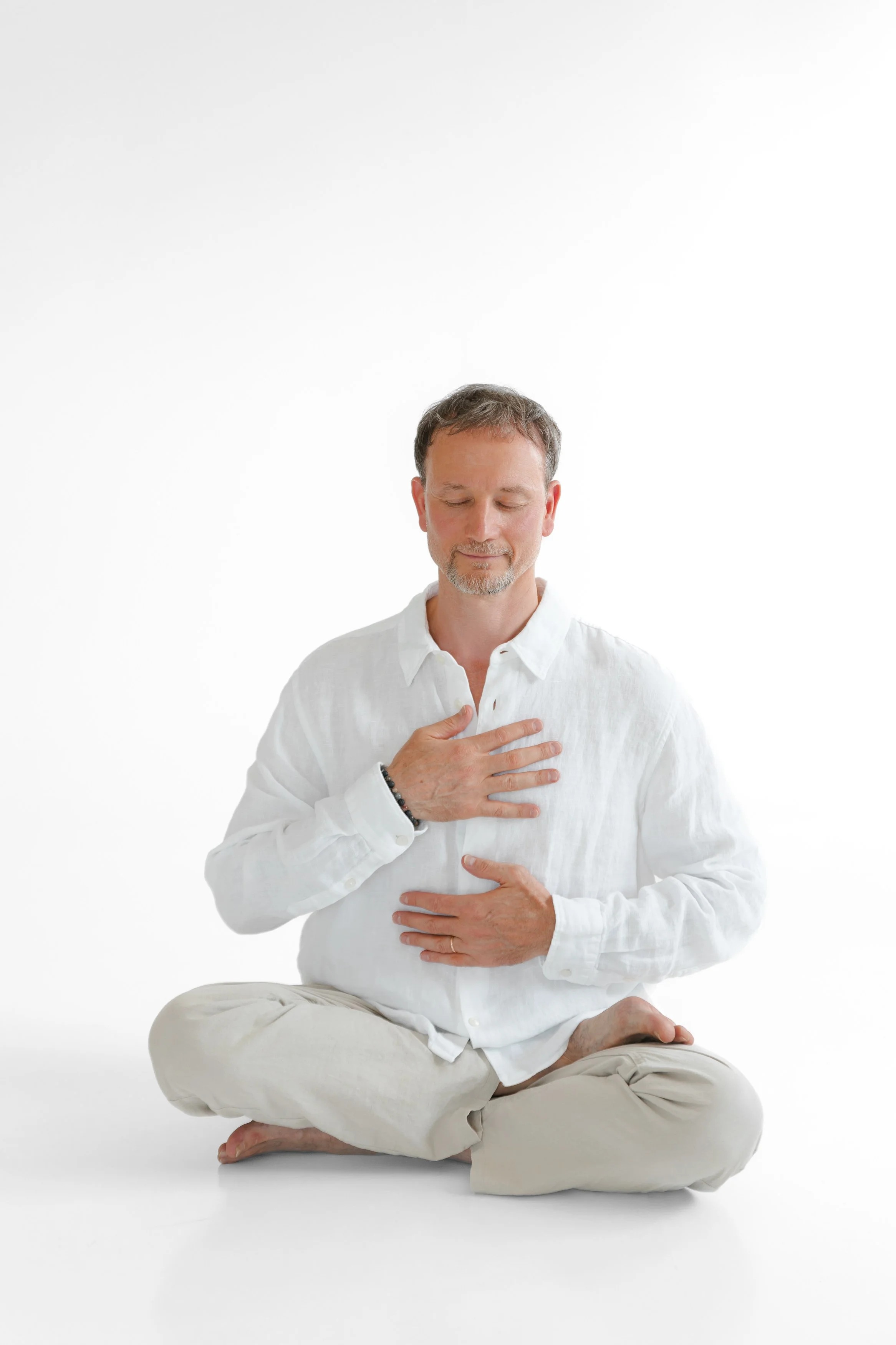 A man with short brown hair and a beard, dressed in a white shirt and beige pants, sitting cross-legged on the floor with eyes closed, holding his left hand on his chest and his right hand on his stomach in a meditative pose.
