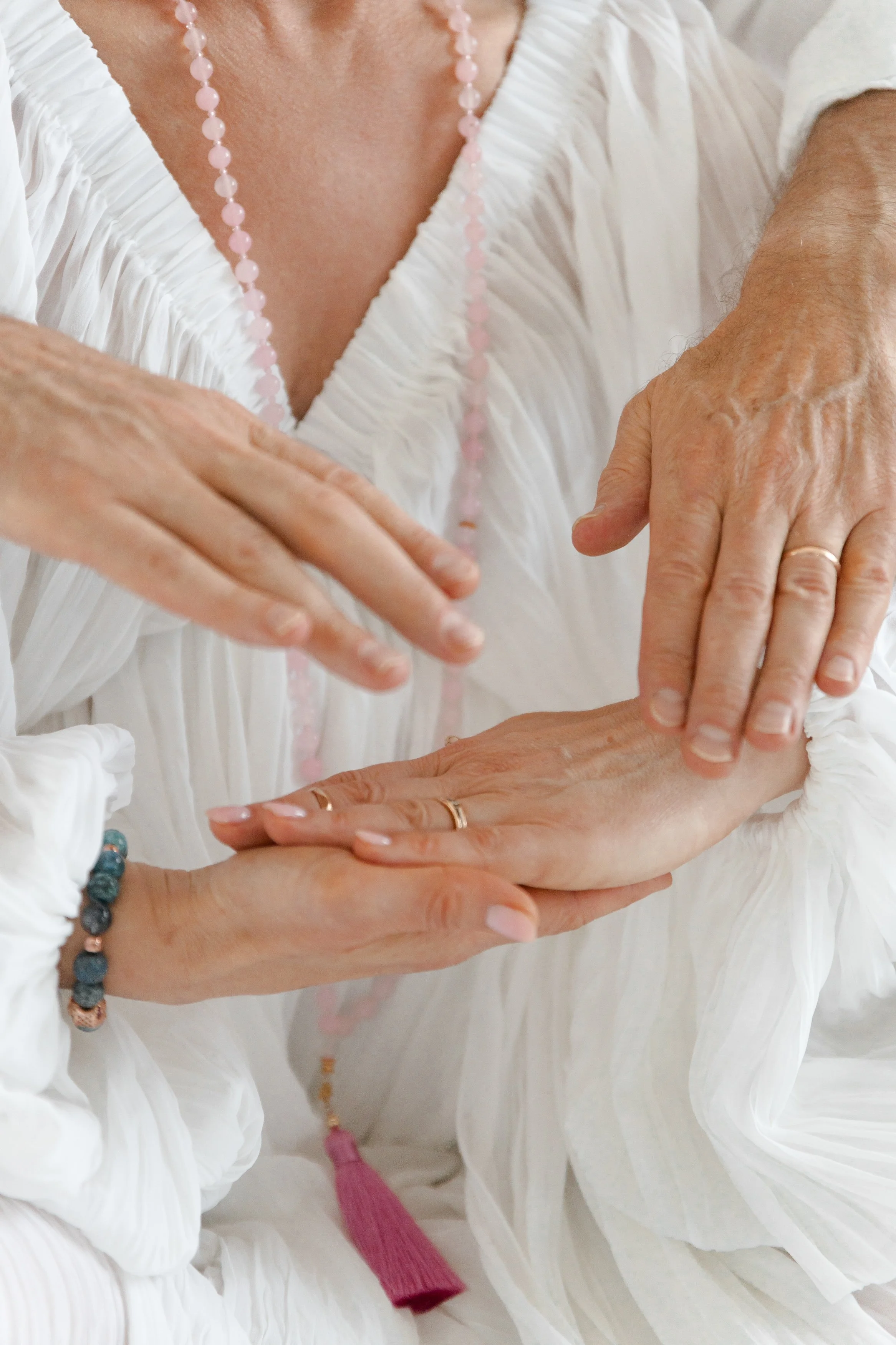 Close-up of an elderly woman with her hands resting on top of each other, wearing rings and bracelets, dressed in a white outfit with pink beaded jewelry.