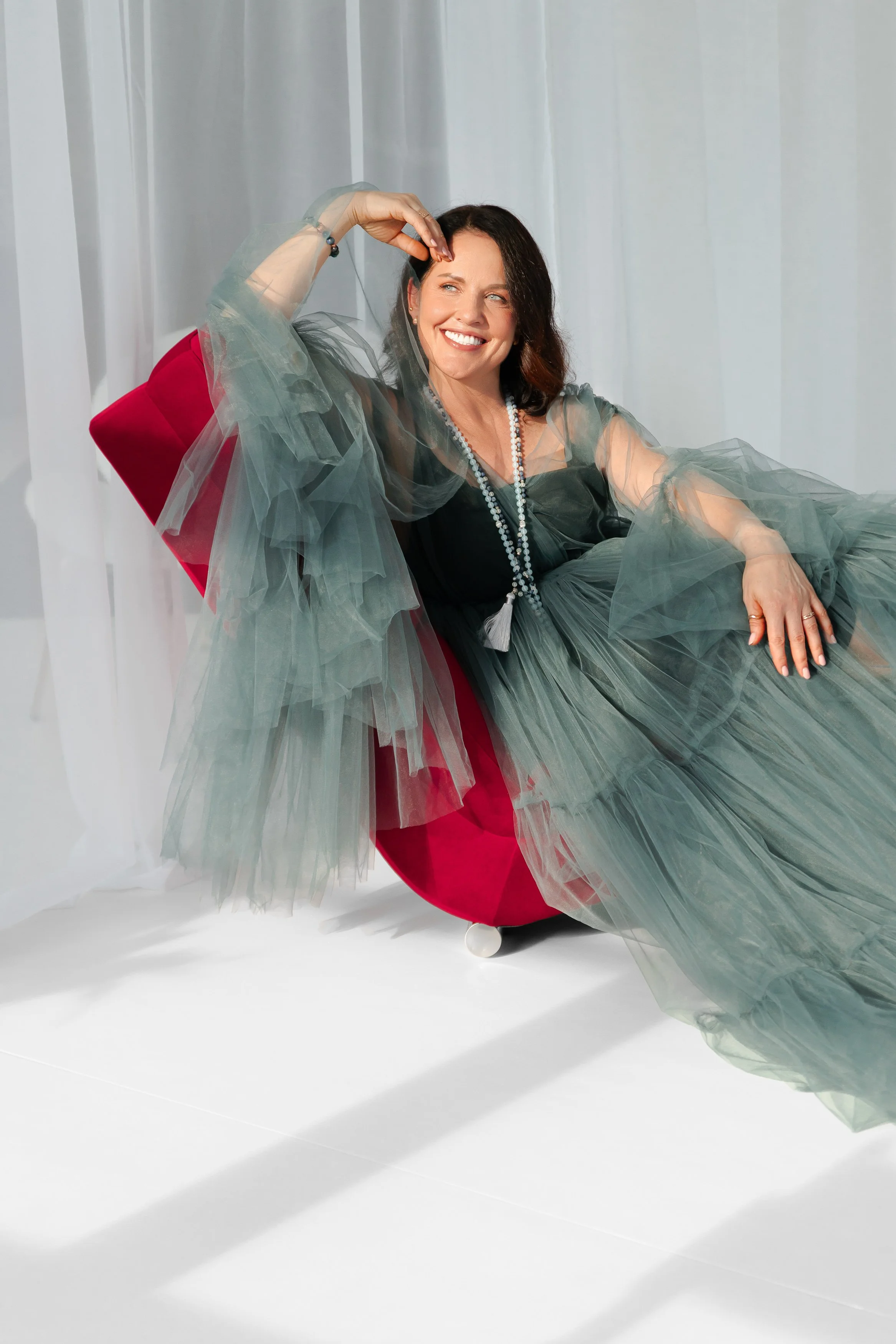 A woman with dark shoulder-length hair sitting comfortably on a red velvet swivel chair, wearing a black dress with sheer green ruffled layers. She is smiling, with one hand on her head and the other on her lap, in front of a white background with vertical sheer curtains.