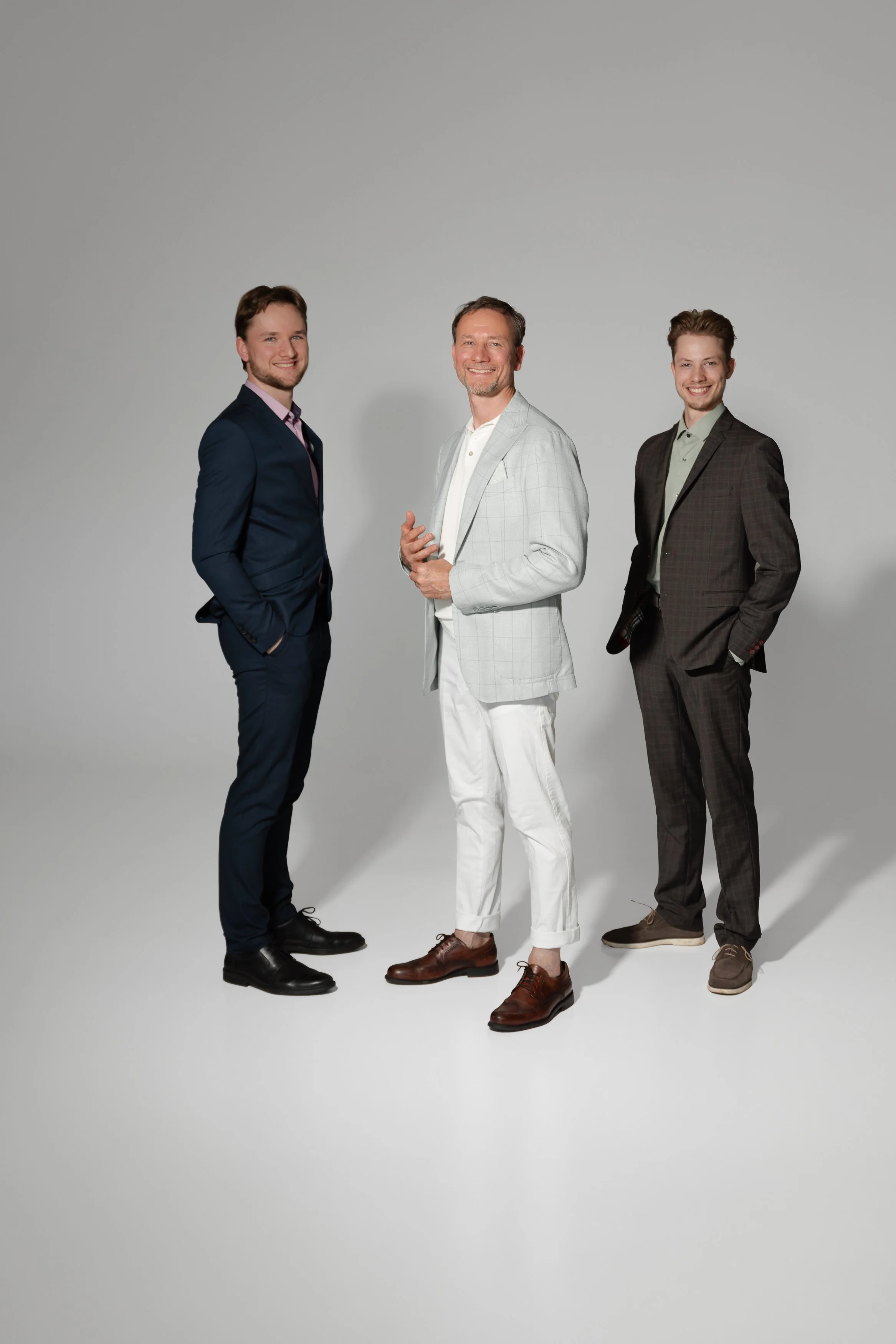Three men in suits standing together against a light gray background, smiling at the camera.