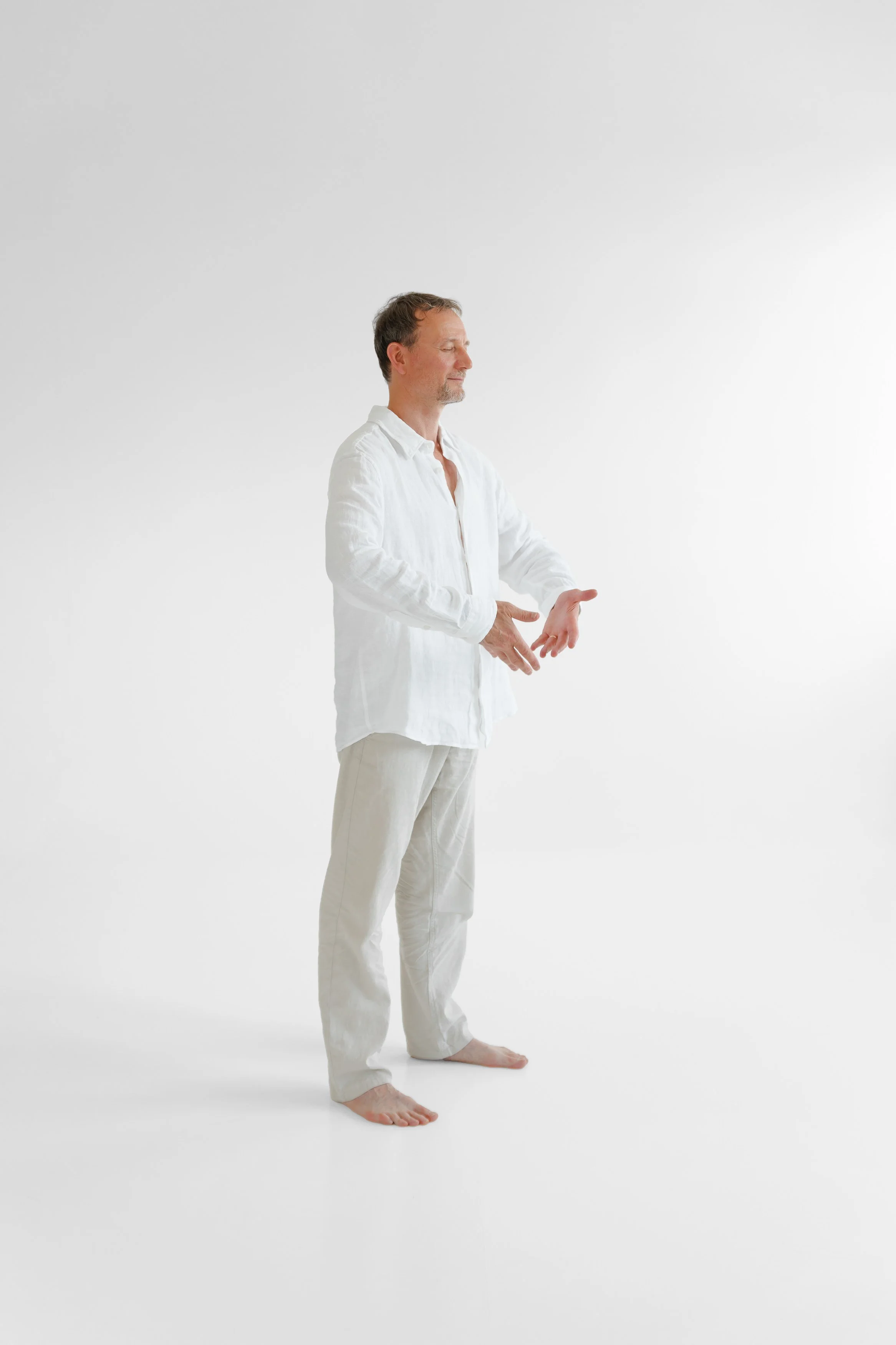 A middle-aged man standing barefoot in a white room, wearing a white shirt and light-colored pants, with his eyes closed and hands in a relaxed position.