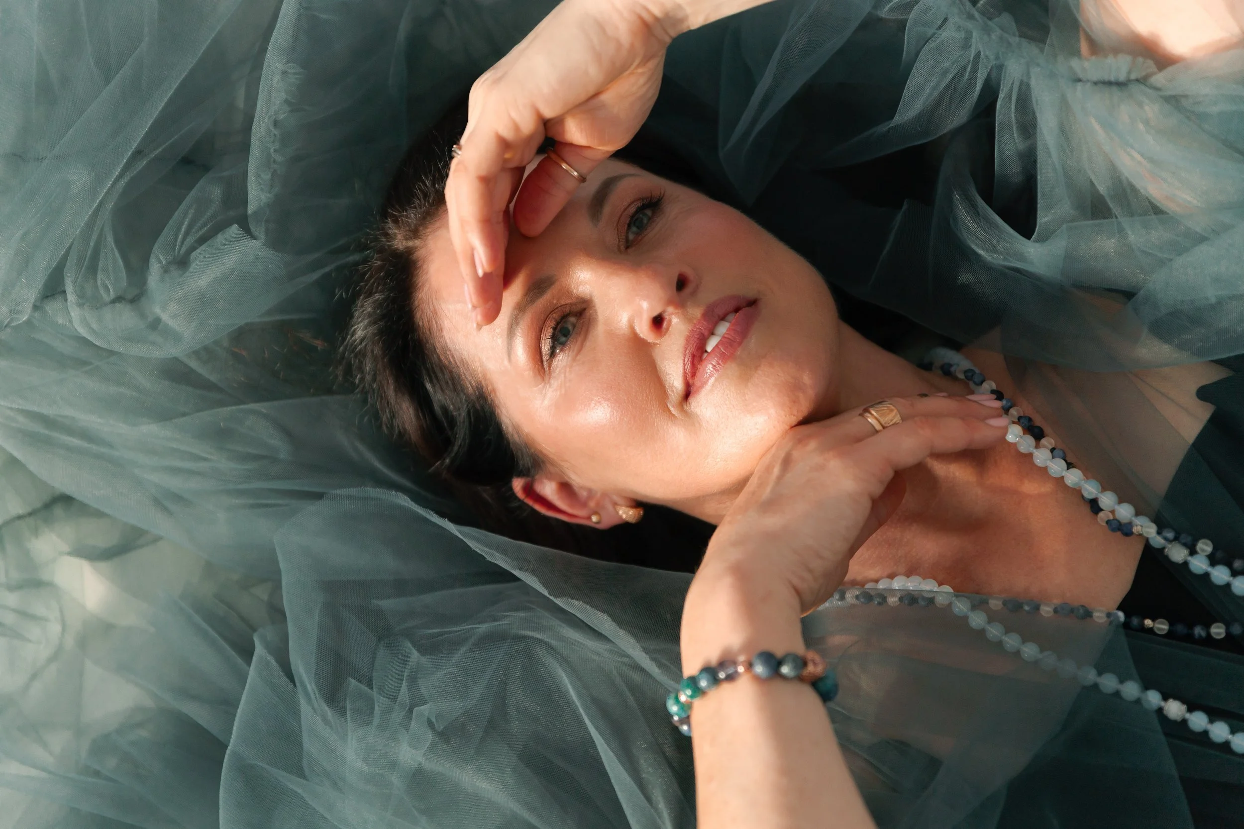 A woman lying on a surface surrounded by dark green tulle fabric, wearing a necklace and bracelets, with her hand resting on her face and shoulder, and looking directly at the camera.