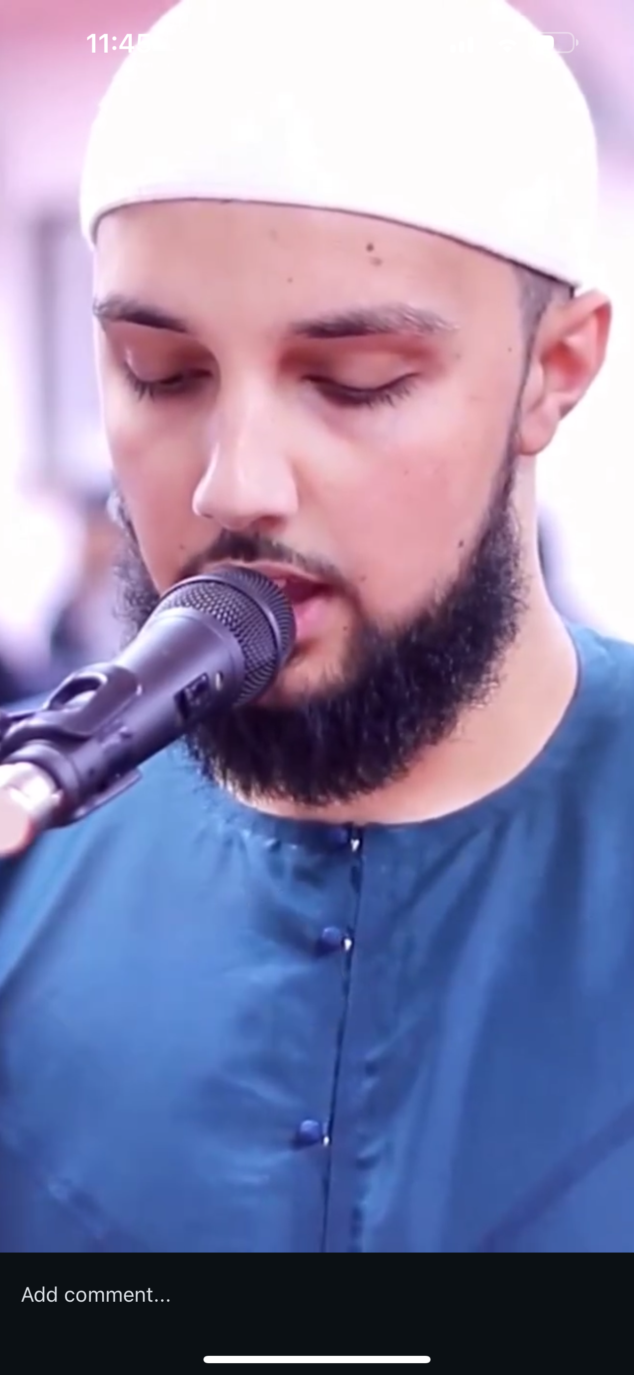 A man with a beard and closed eyes wearing a white cap and dark clothing, speaking into a microphone.