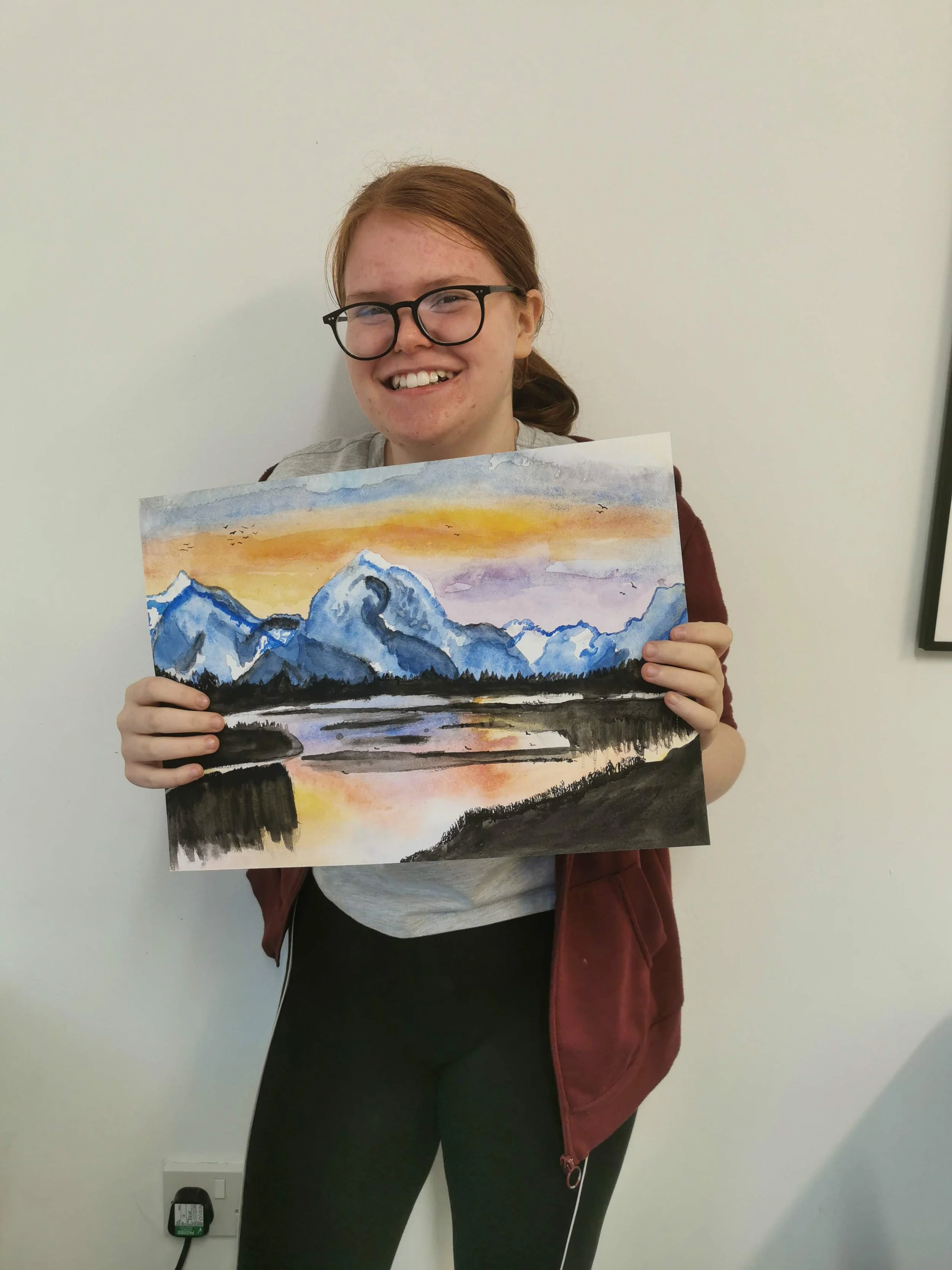 A girl with glasses holds a landscape watercolor painting of mountains, a river, and a colorful sunset.