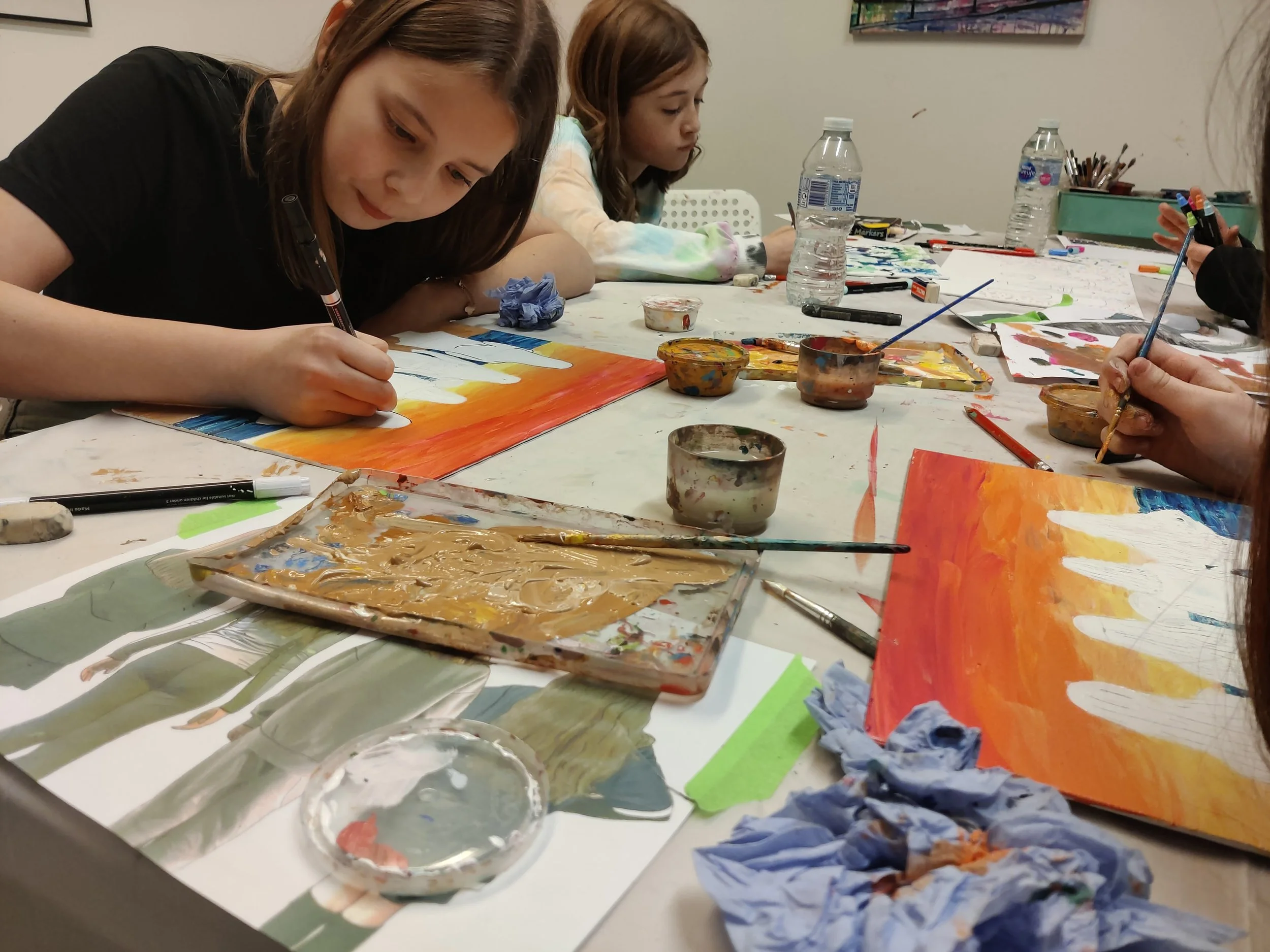 Three girls painting on canvases at a table with art supplies, water bottles, and crumpled tissues.