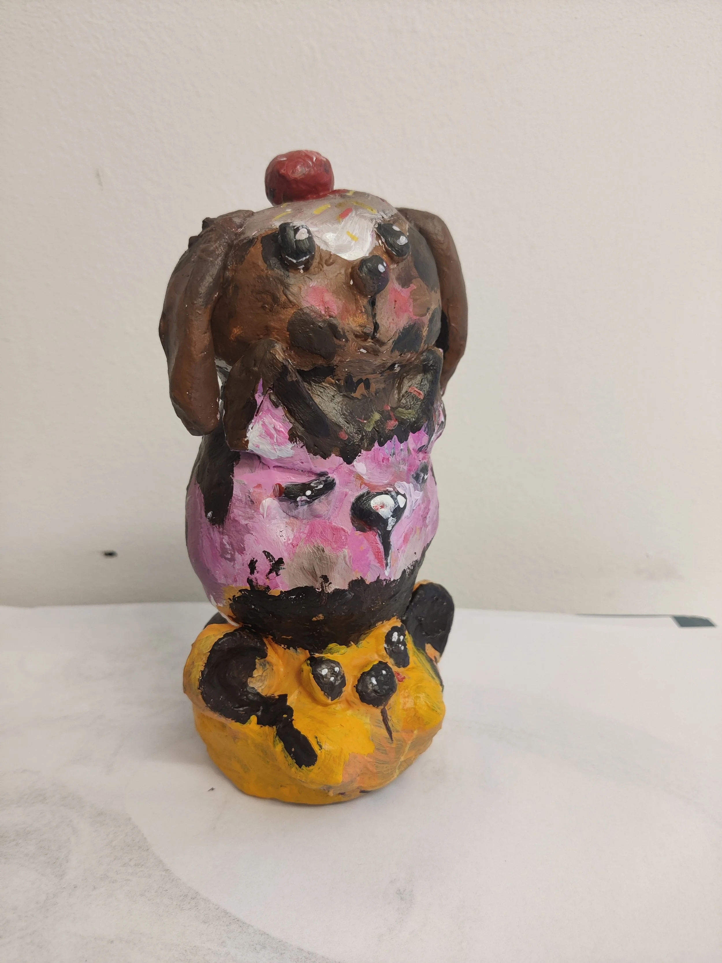 Hand-painted sculpture of a cute dog with floppy ears, wearing a pink dress and a yellow skirt with black spots, placed against a plain background.