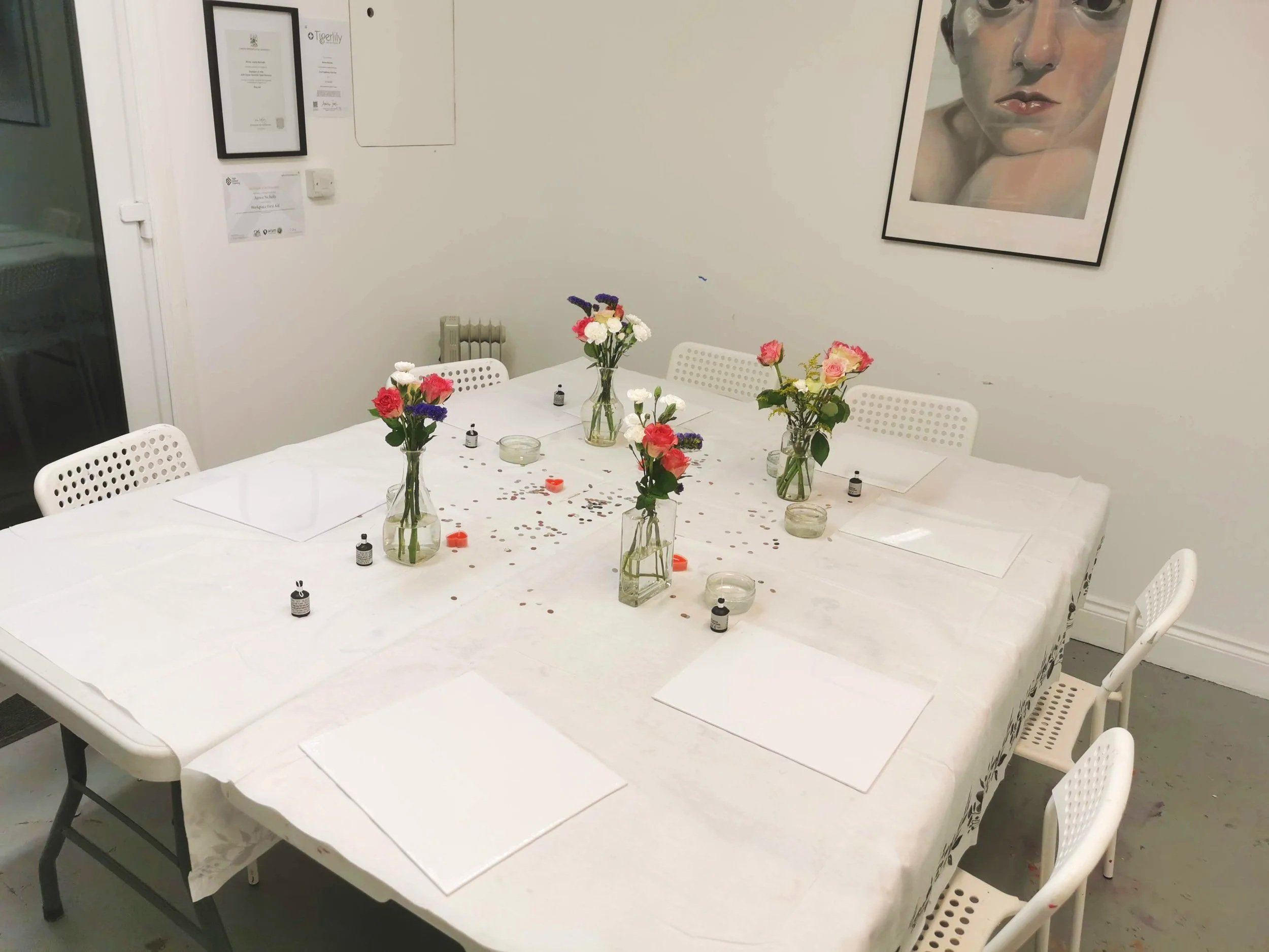 A white rectangular table set with five glass vases filled with pink, white, and purple flowers. Scattered around the vases are small bottles of essential oils, candles, and confetti. Four white chairs surround the table, with four white placemats set around it. On the wall, there is a large portrait of a face and some certificates or artwork framed on the wall.