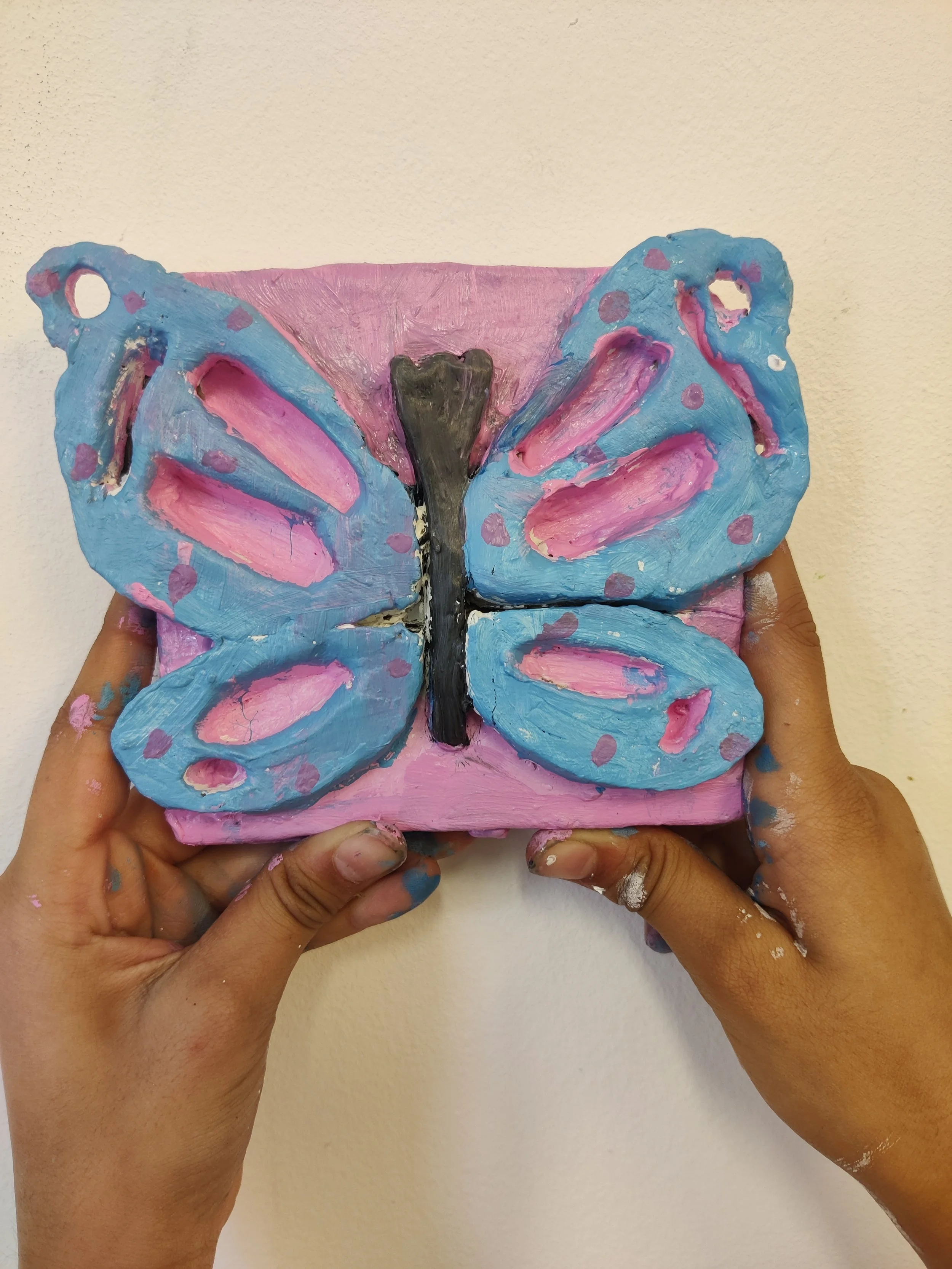 Child's hands holding a pink square block with a blue butterfly painted on it, wings with pink spots and black body, against a plain beige wall.