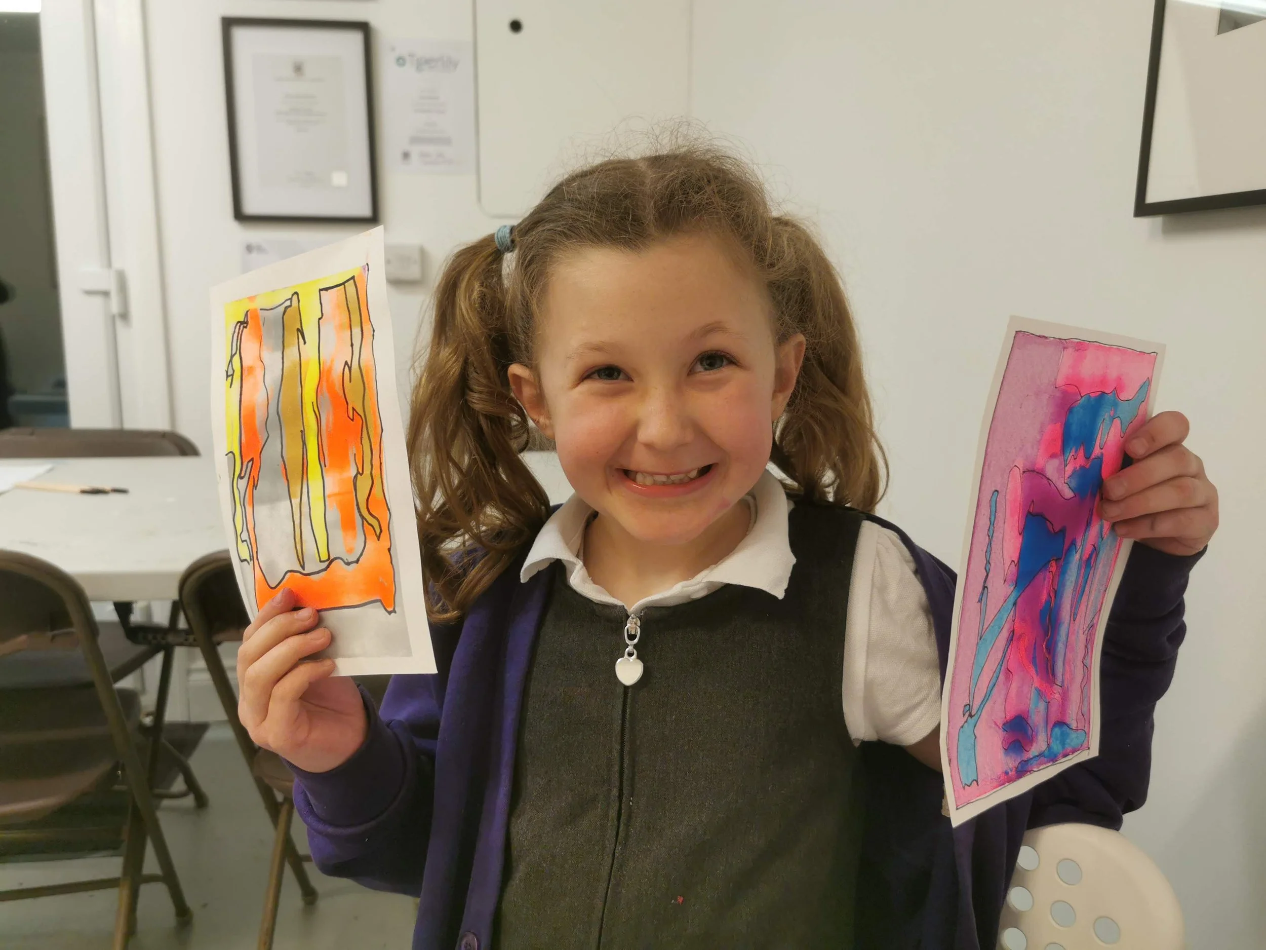 A girl with curly hair holding up two colorful pieces of artwork, smiling at the camera.