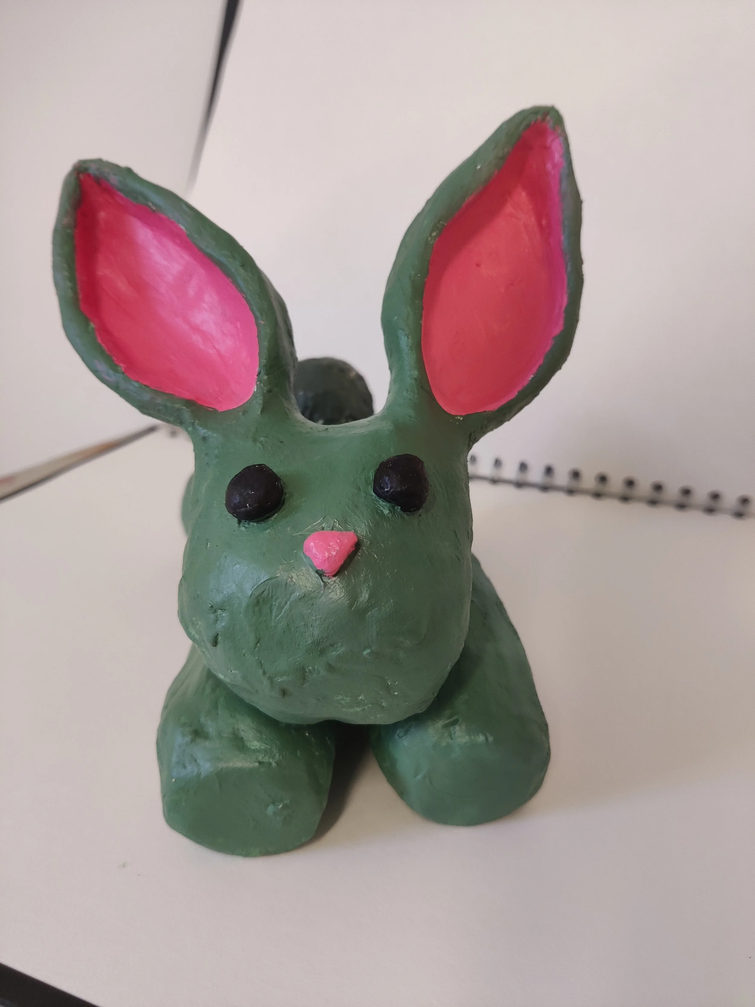 Handmade clay sculpture of a bunny painted in green with pink inner ears, pink nose, and black eyes, placed on a white surface.