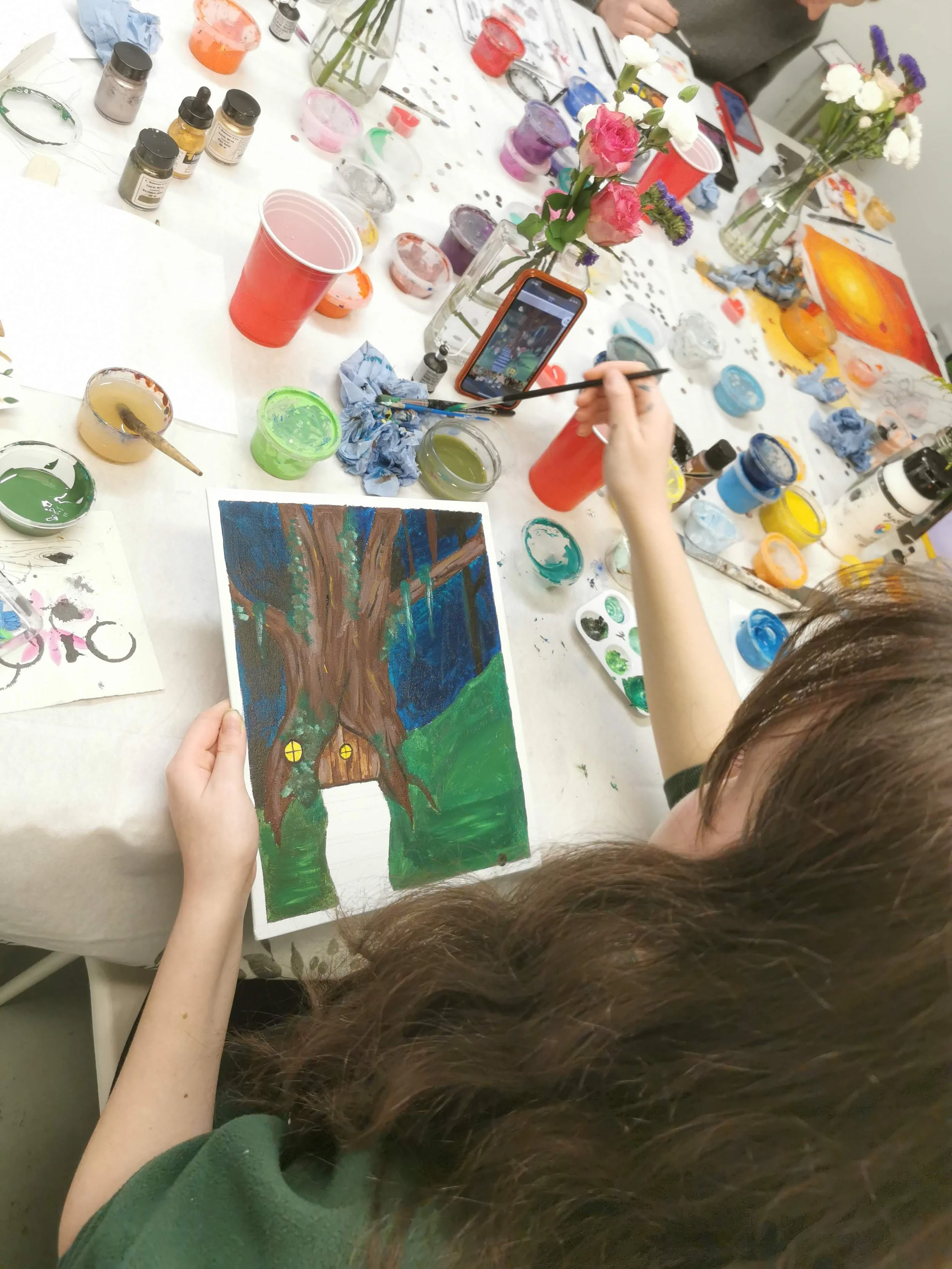 A person painting a picture of a large tree with a small cottage at its base, using vibrant colors. The workspace is filled with various paints, brushes, and art supplies, and the person is holding a cup and a paintbrush, working on the artwork surrounded by flowers and art materials.