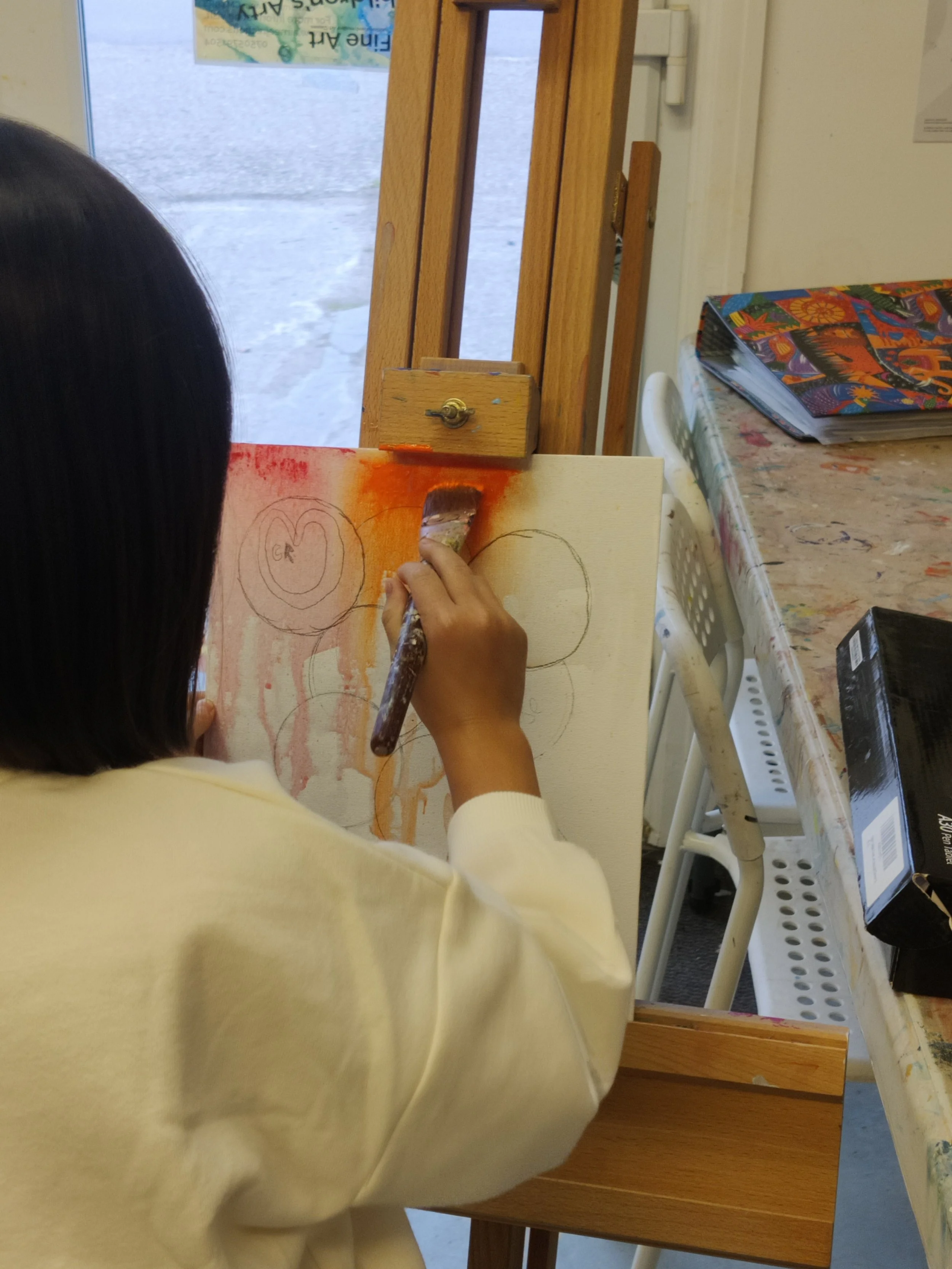 A child with dark hair and wearing a light-colored shirt paints on a canvas using watercolors. The painting appears to have circles with some orange and red paint around them. The child is sitting at a cluttered table with art supplies and a colorful