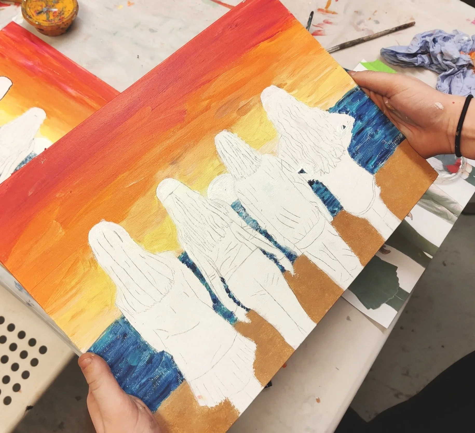 A person holding a painting of four girls with wings walking on a beach during sunset, with the sky painted in warm tones of orange, yellow, and red. The girls are outlined in white without detailed features.