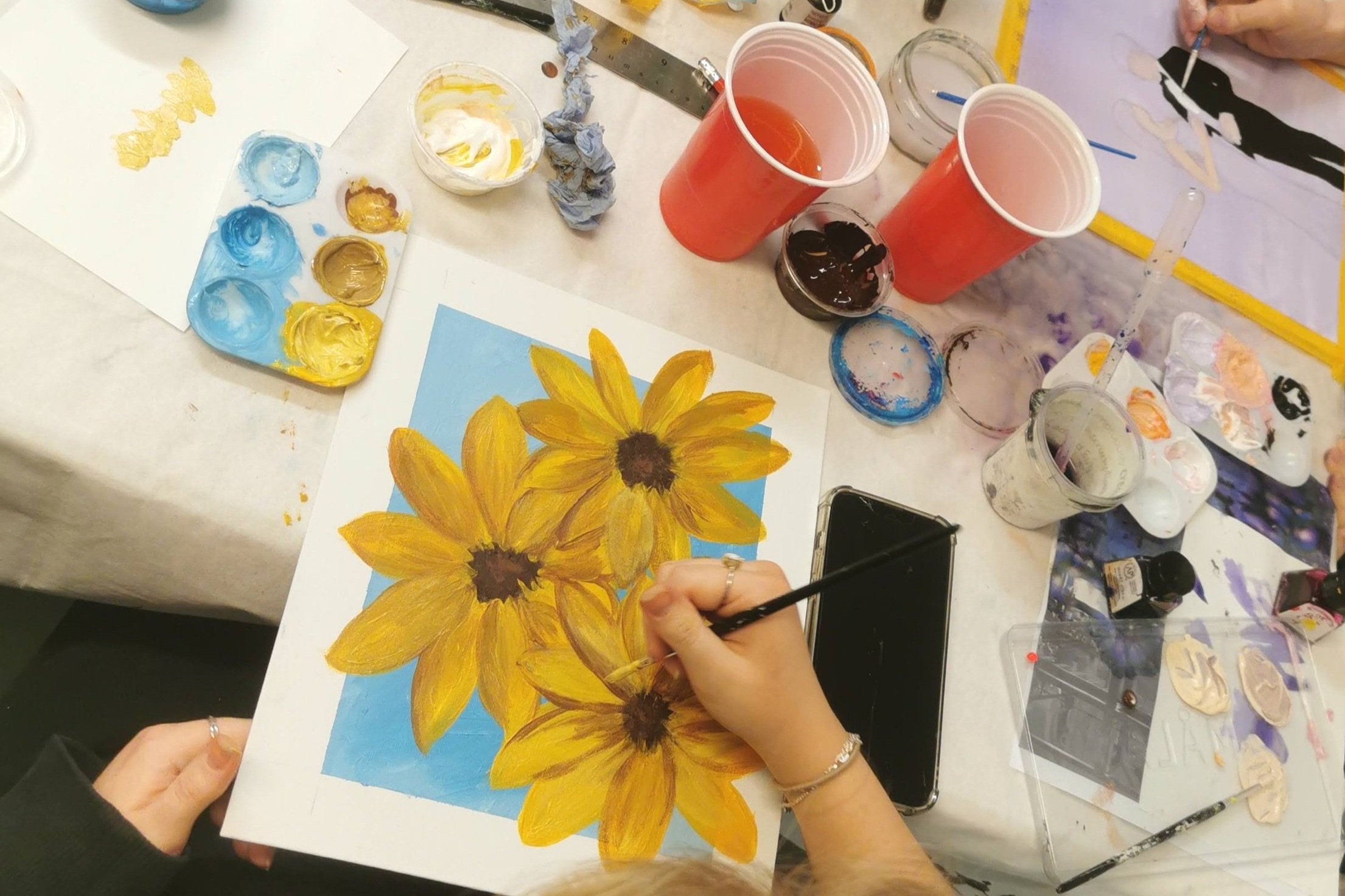 A person painting a floral artwork of yellow sunflowers on a piece of paper, surrounded by various painting supplies, cups of beverage, and paint palettes on a table.