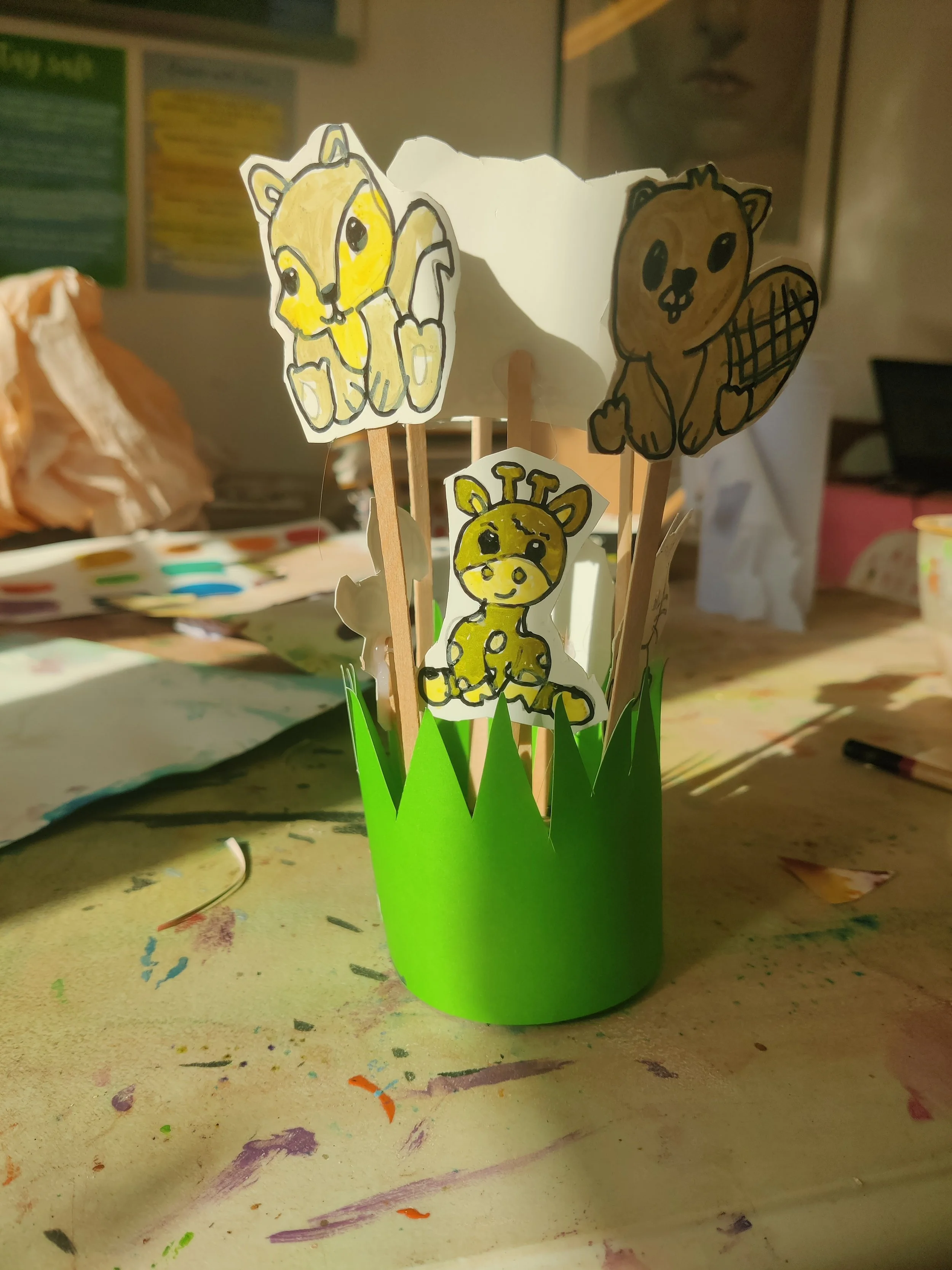 A paper craft featuring cartoon animals, including a lion, giraffe, and bear, attached to sticks and placed in a green paper grass container.