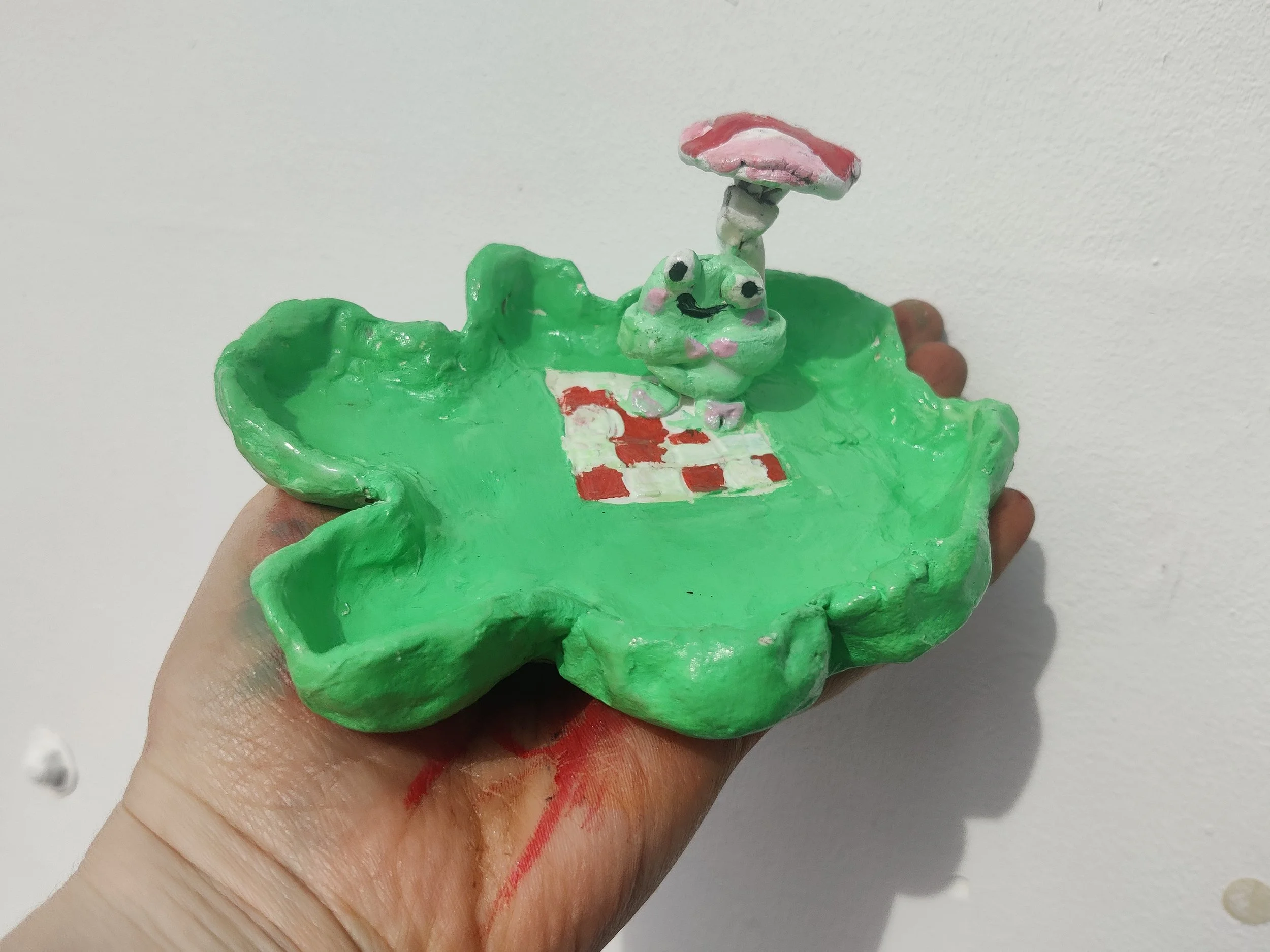 A handmade clay art piece of a frog holding an umbrella standing on a lily pad with a checkered pattern.