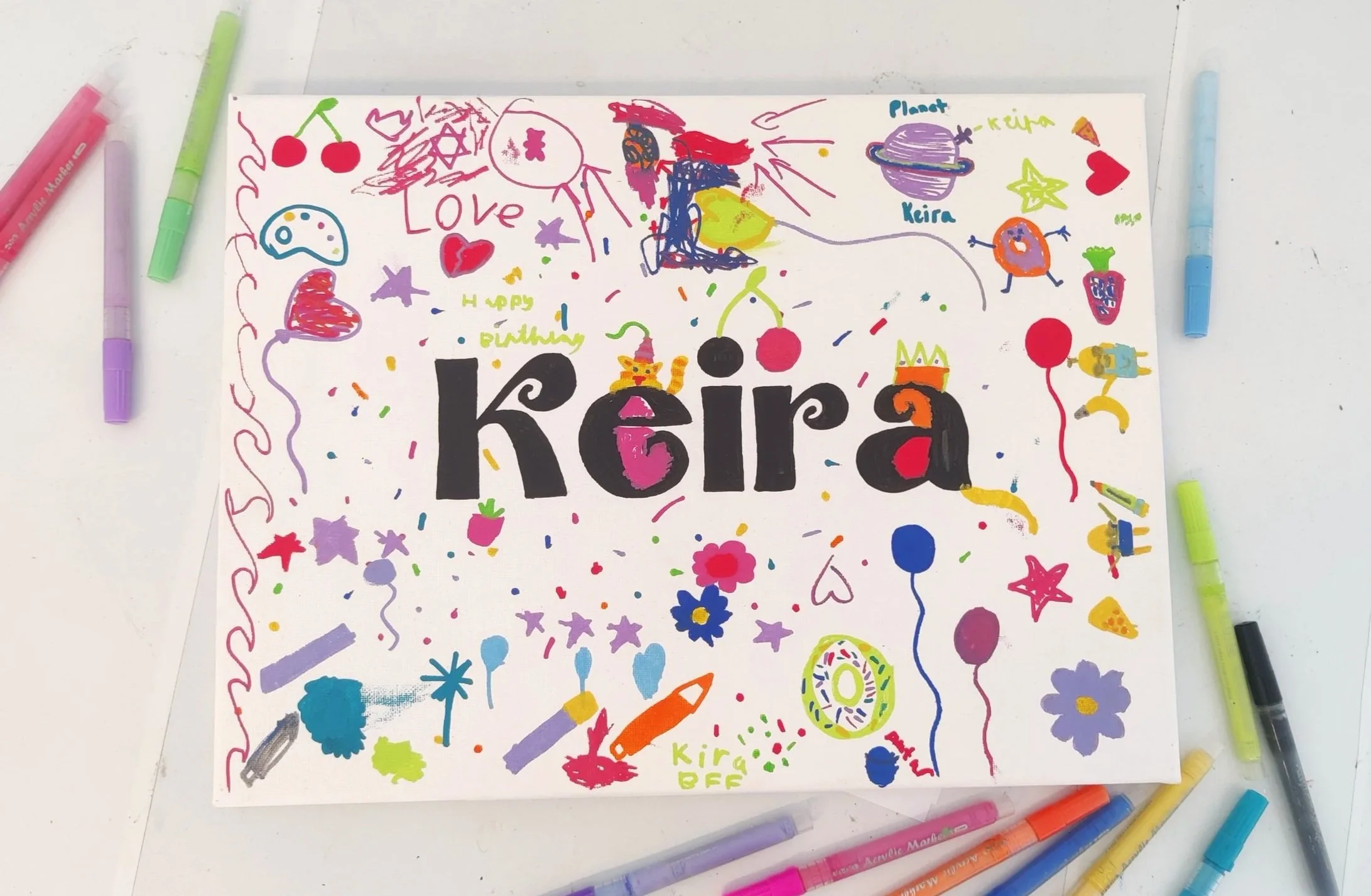 Colorful hand-drawn doodles and doodles surround the name 'Kaira' on a white paper, with various drawings of hearts, cherries, planets, stars, flowers, and other whimsical designs in different colors.