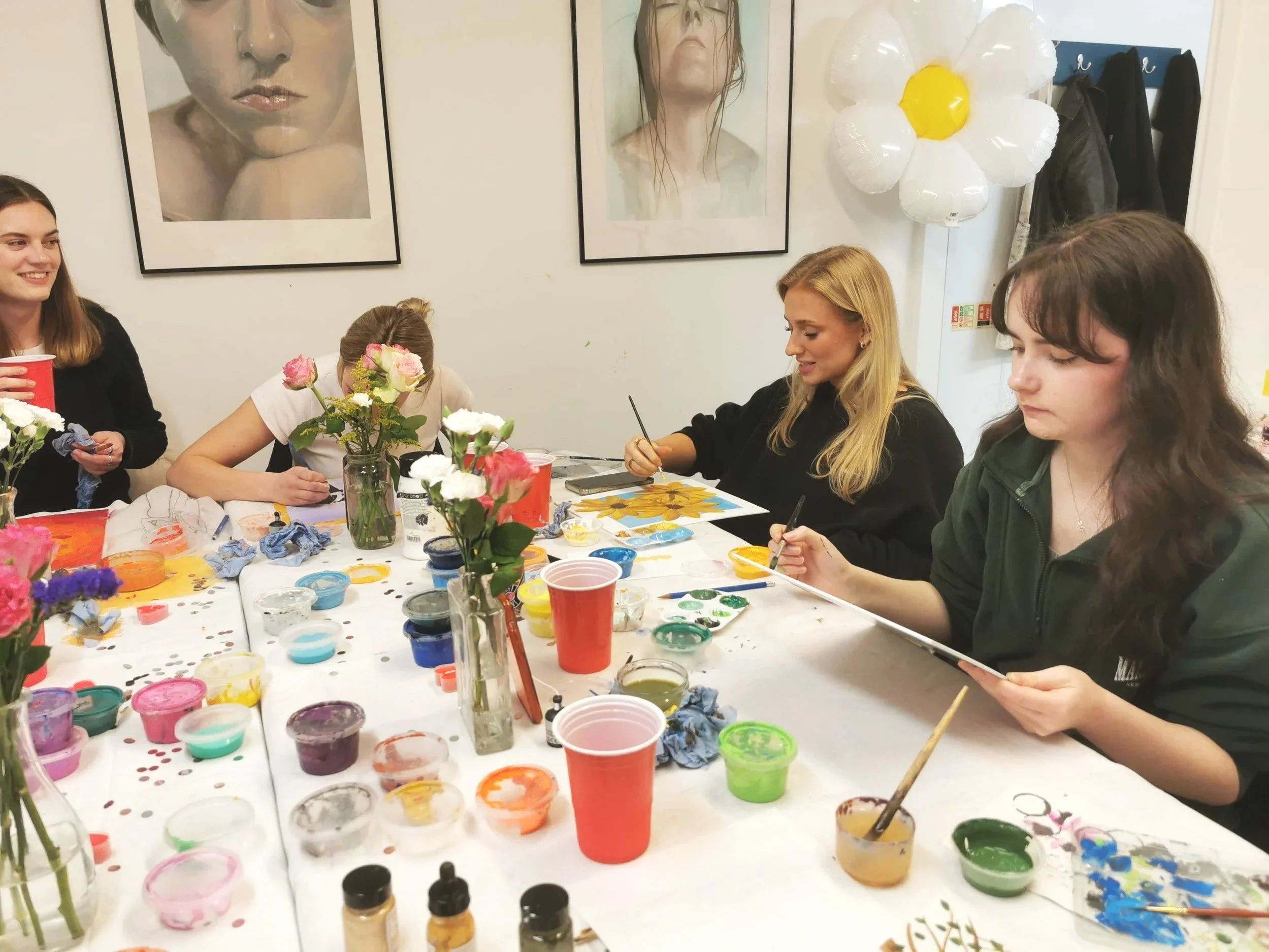 Group of four women painting and creating flower-themed art projects at a table covered with painting supplies, including paints, brushes, and cups, in an art studio with large floral paintings on the wall.