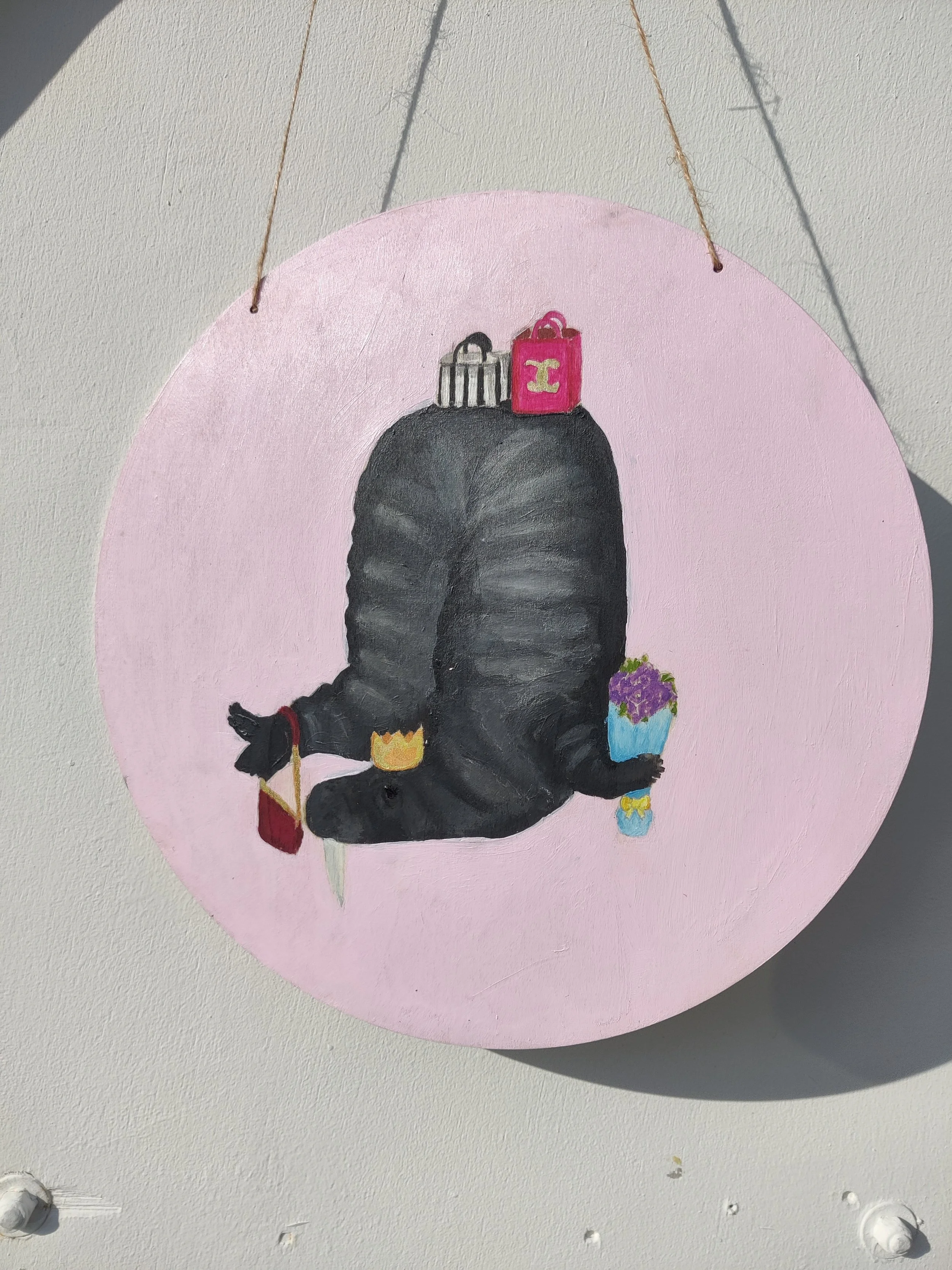 Painting of a black animal, possibly a mole or rat, lying on its back with various small objects on and around it, including a crown, a shopping bag, a flower pot, and small containers, with a light pink circular background.