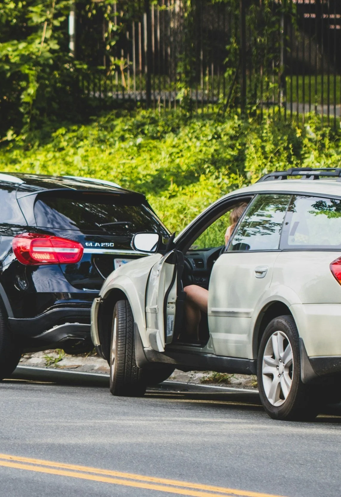 Zwei Autos, die in einen Unfall verwickelt sind, mit einer Person im beigefarbenen Auto. Der schwarze Wagen ist hinten beschädigt, die Autos stehen auf einer Straße mit gelber Linie. Im Hintergrund grüne Bäume und eine Metallzäune.