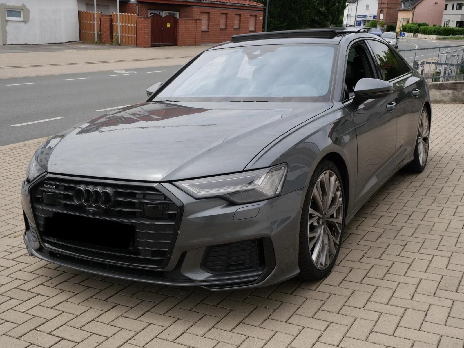 Ein grauer Audi A5 Limousine, parkt auf einem gepflasterten Parkplatz neben einer Straße in einem Wohngebiet.