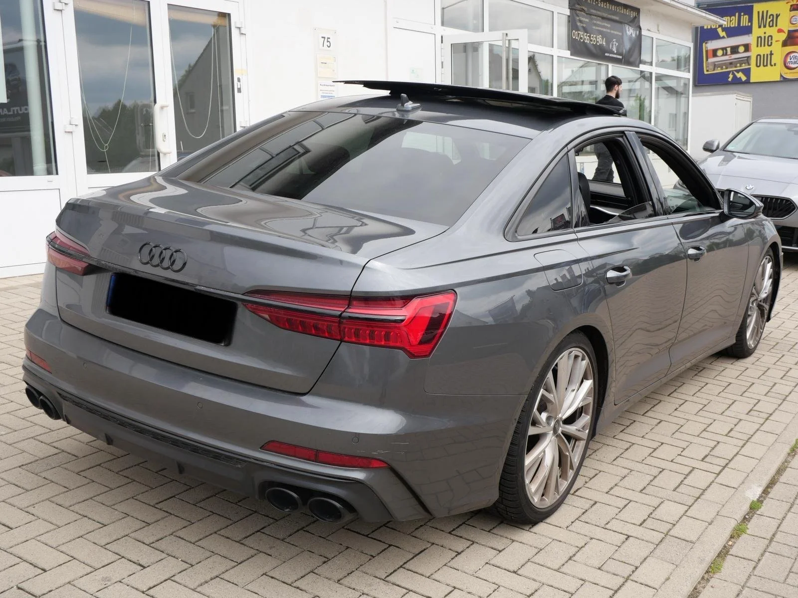 Grauer Audi-Auto parkt vor einem Gebäude mit Glasfenstern und mehreren anderen Autos im Hintergrund.