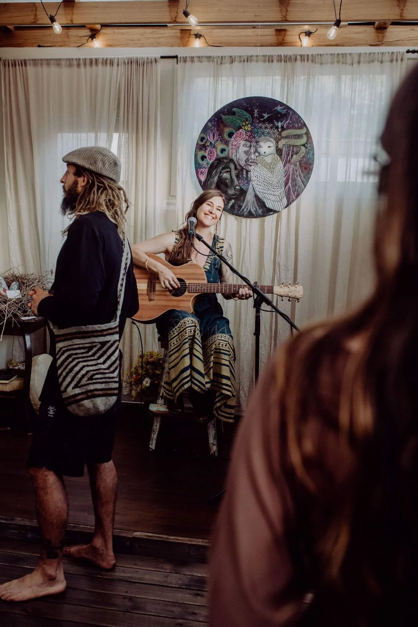 A woman playing acoustic guitar and singing into a microphone during a performance, with a colorful circular artwork on the wall behind her. A man with dreadlocks and a cap stands nearby, and audience members are visible in the foreground.