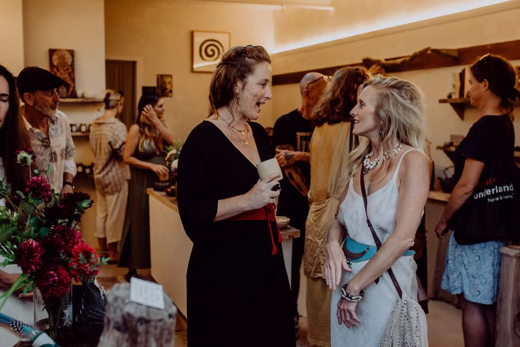 Two women talking and smiling at an indoor social gathering, with other guests in the background.