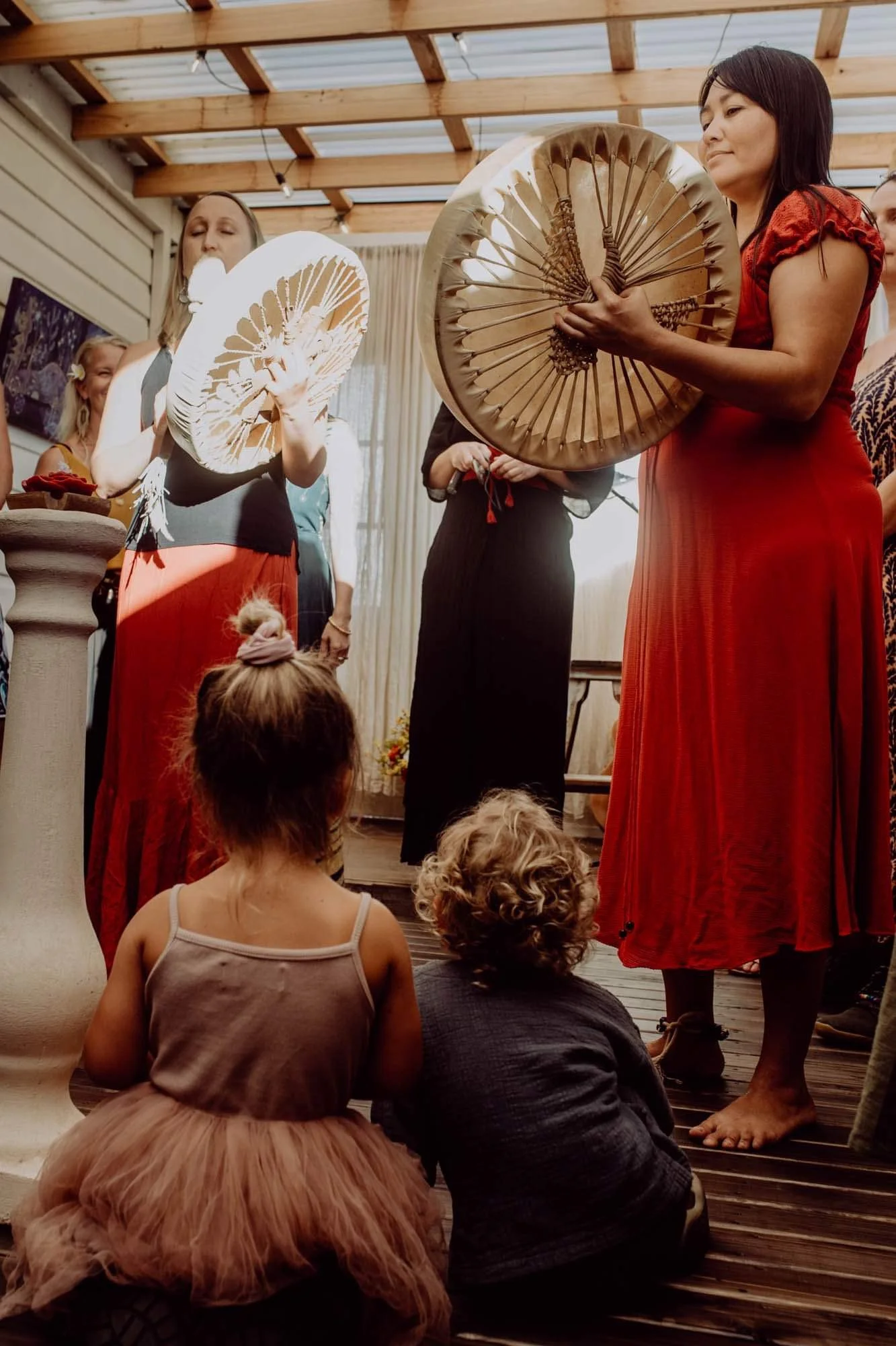 People holding large fans during a cultural or artistic performance inside a wooden-roof venue. Two children are sitting on the floor observing the event.