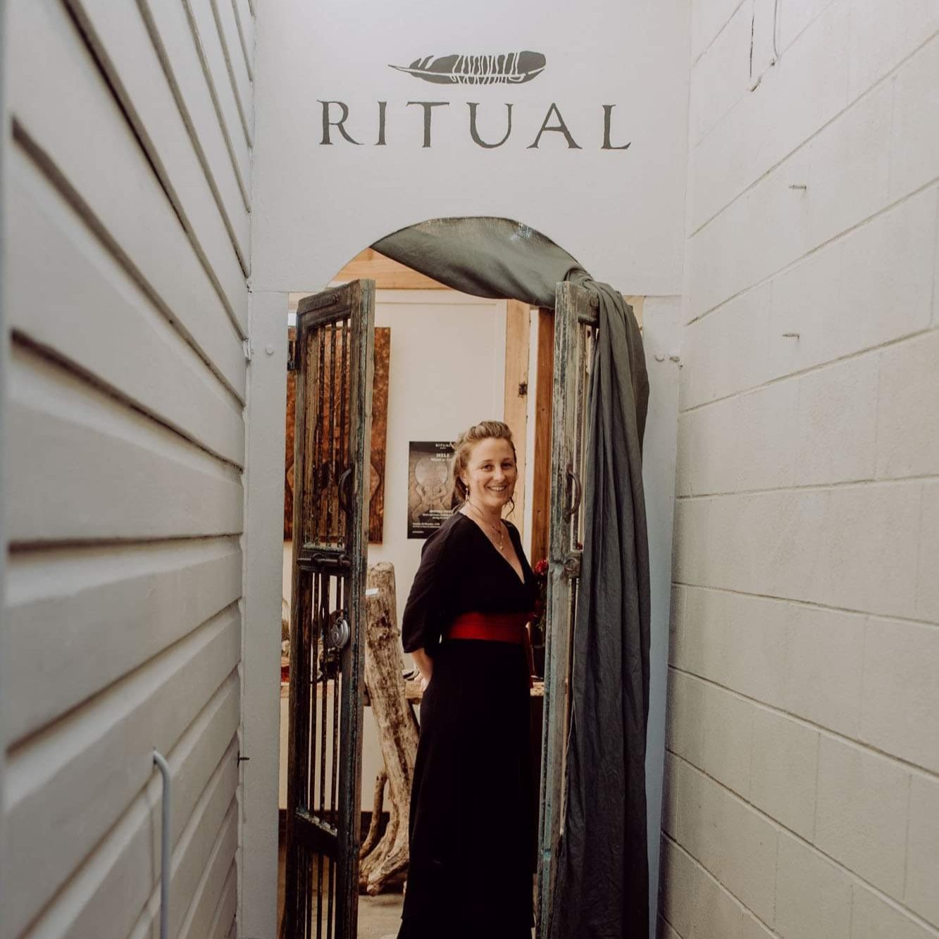 Woman smiling at the entrance to a ritual space, standing inside a small, rustic doorway with a sign above that reads 'RITUAL' and a feather illustration.