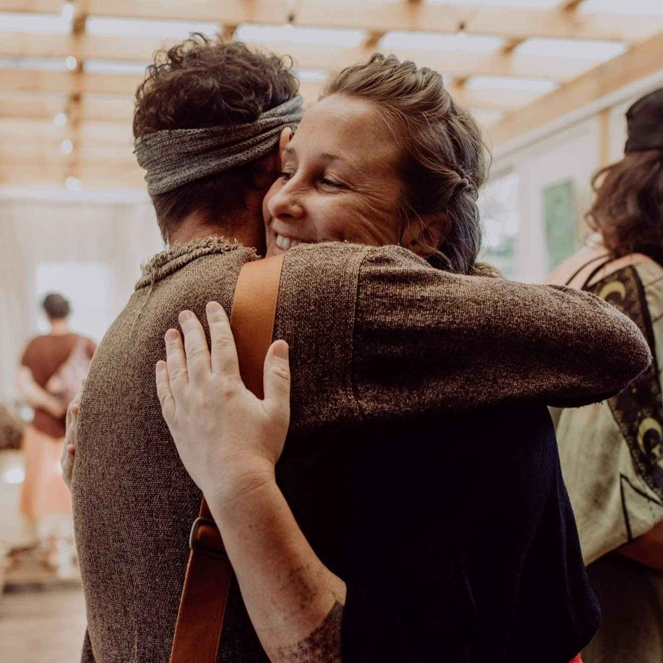 Two women hugging warmly, smiling, in a cozy, light-filled room with people in the background.