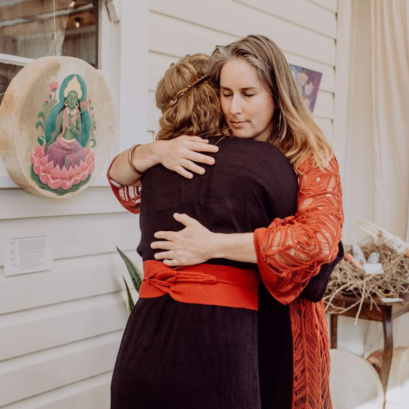Two women embracing, one with brown hair and the other with red hair, in an art gallery with a colorful painting of a deity in the background.