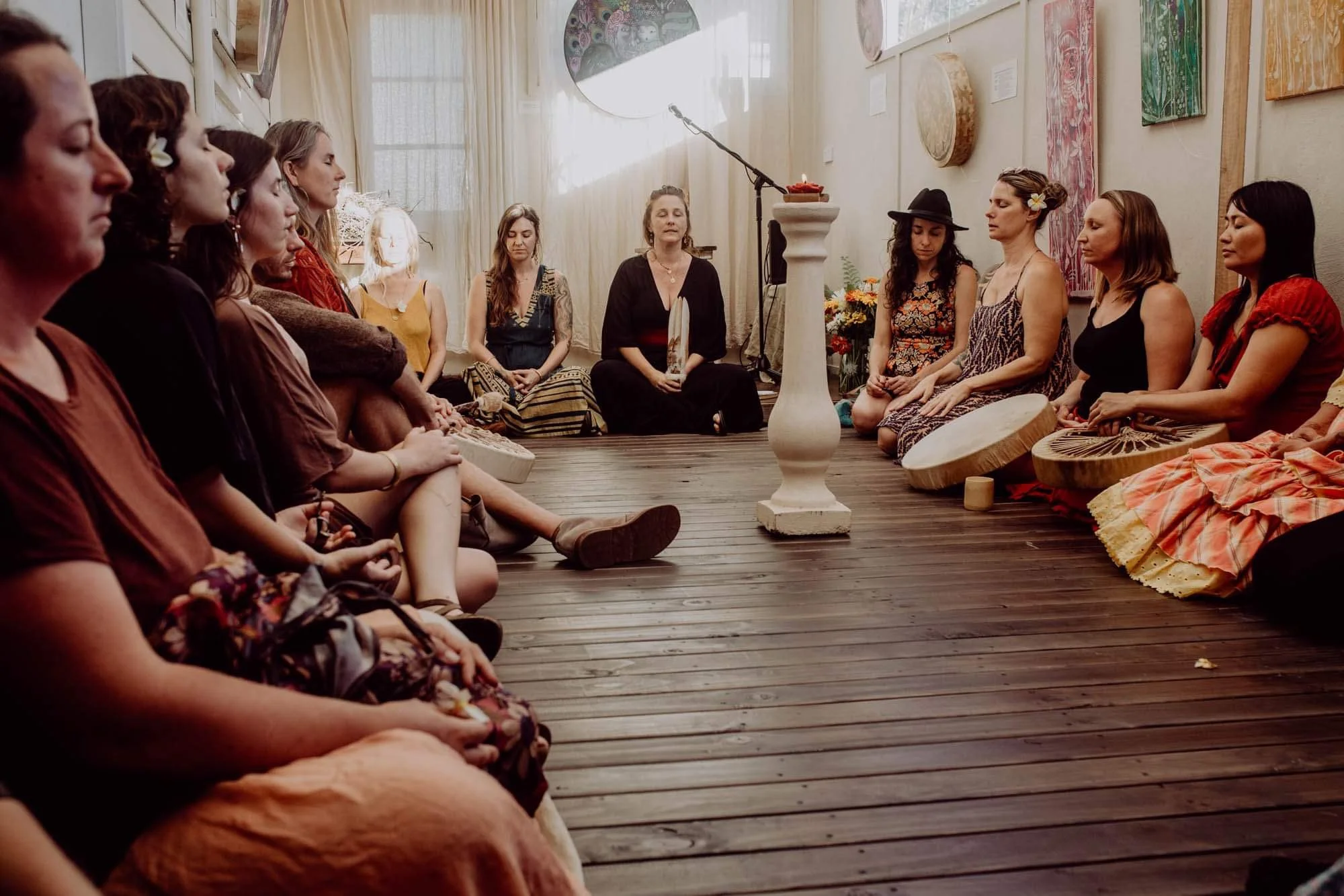 A group of women sitting in a circle on a wooden floor, with some holding drums and one woman at the center with her eyes closed, appearing to meditate or lead a spiritual practice.