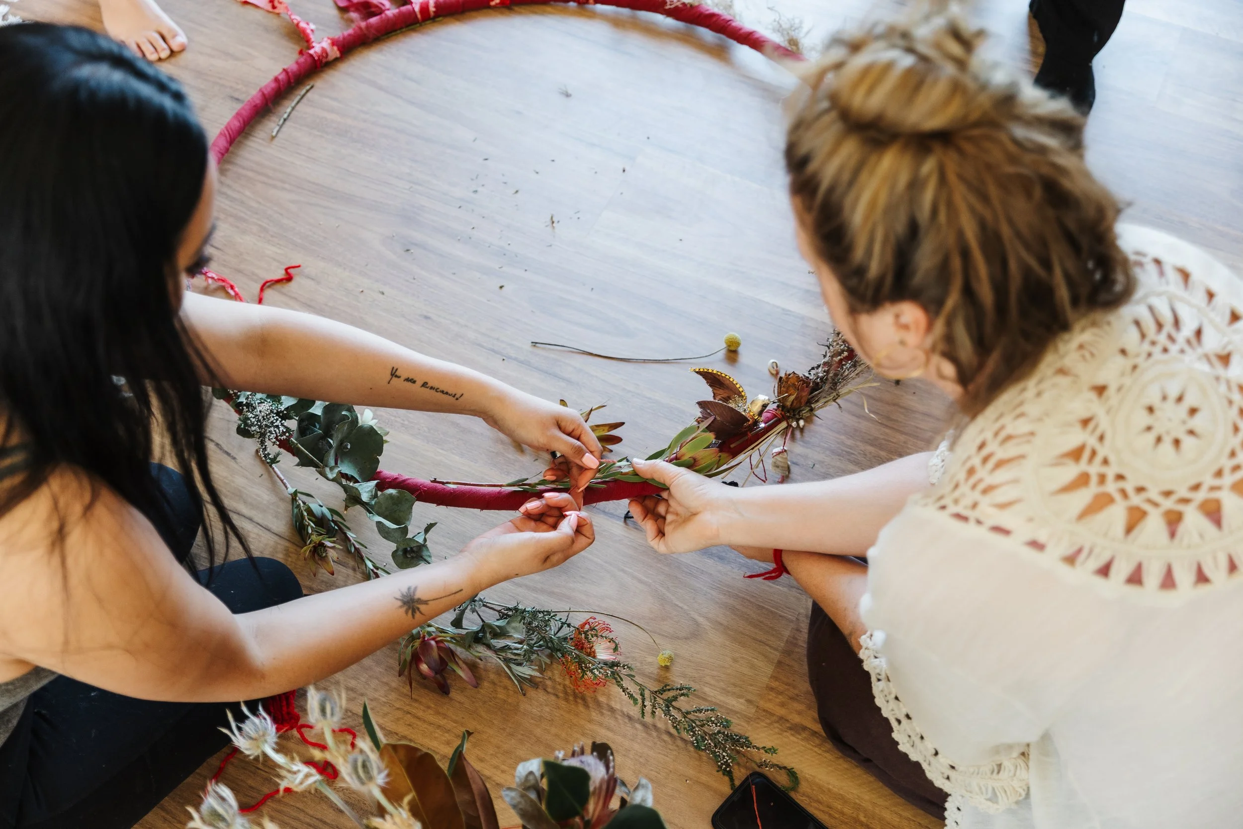 Two women sitting on the floor arranging a floral wreath with flowers, leaves, and branches, on a wooden surface.