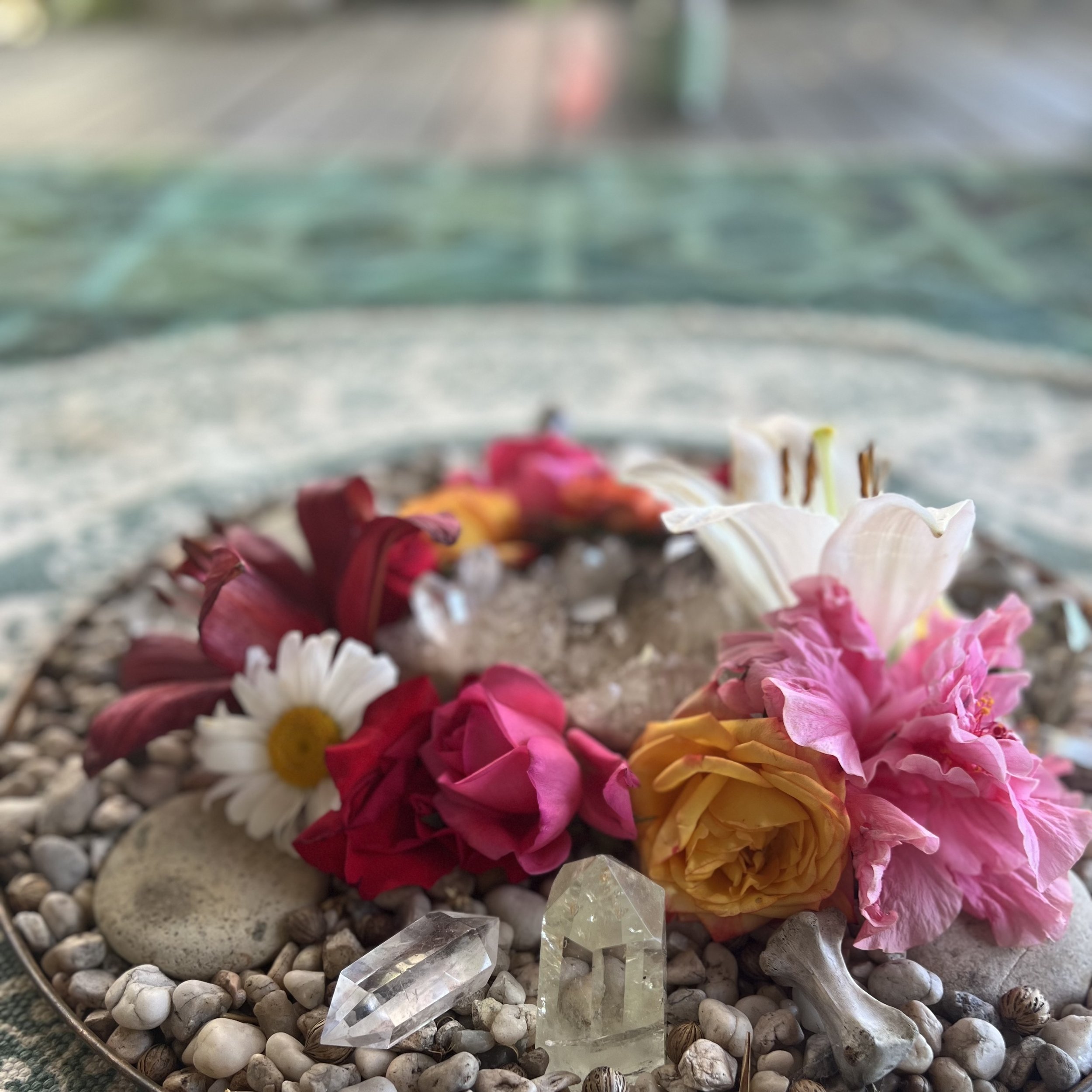A spiritual altar or wreath decorated with colorful flowers, stones, shells, and a clear quartz crystal, set on a bed of pebbles.