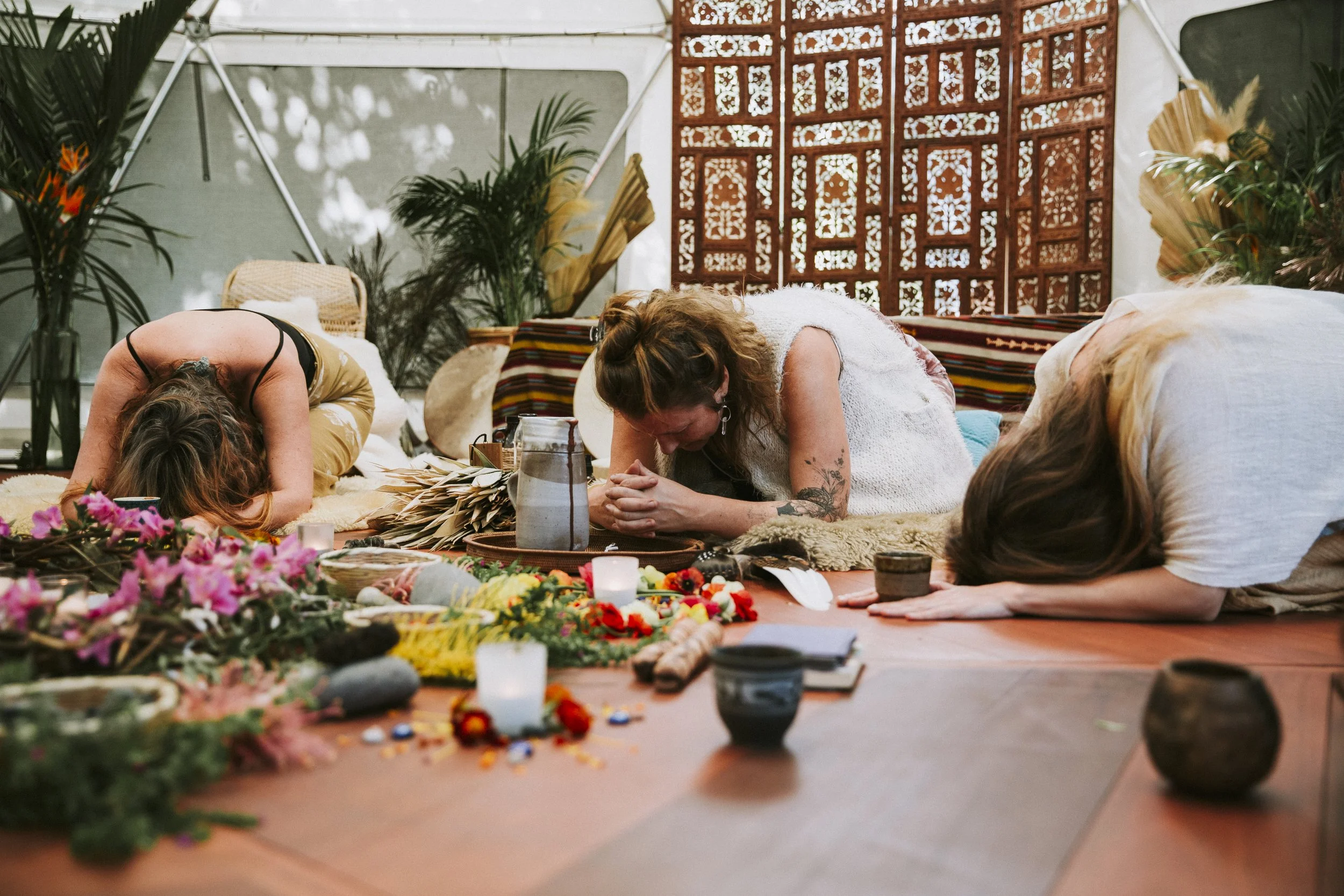 Three women praying or meditating with their heads bowed and hands clasped, surrounded by flowers, candles, and spiritual objects in a peaceful, decorated setting.