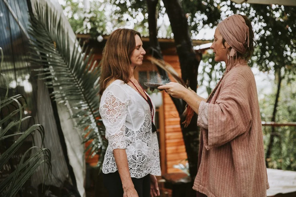 Two women standing outdoors, one offering a plate to the other, with greenery and a wooden structure in the background.