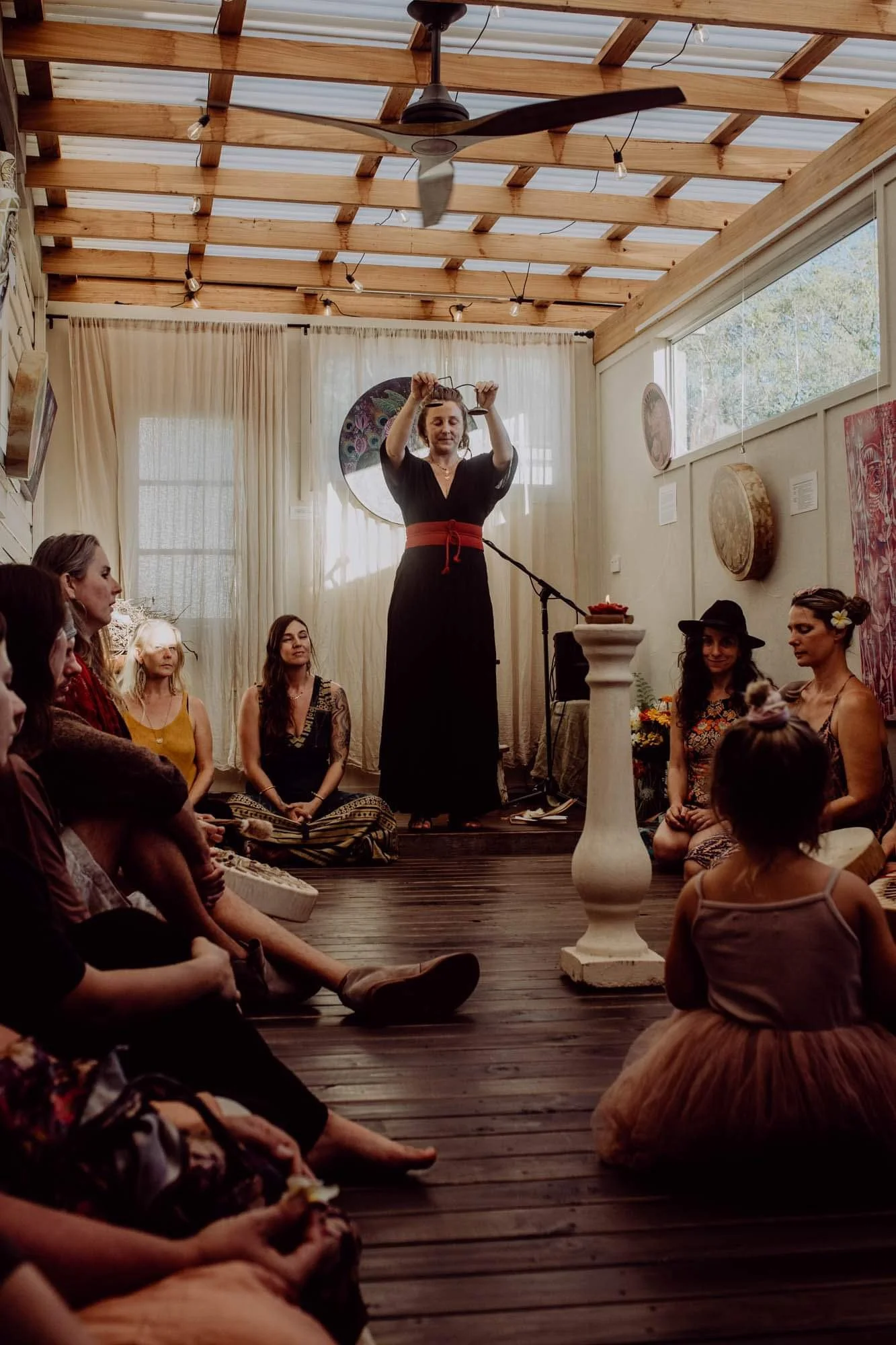 A woman in a black dress with a red sash performs a hoop dance on a stage in front of an audience seated on the wooden floor. The room has a rustic style with wooden ceiling beams, hanging lights, and various artwork on the walls.