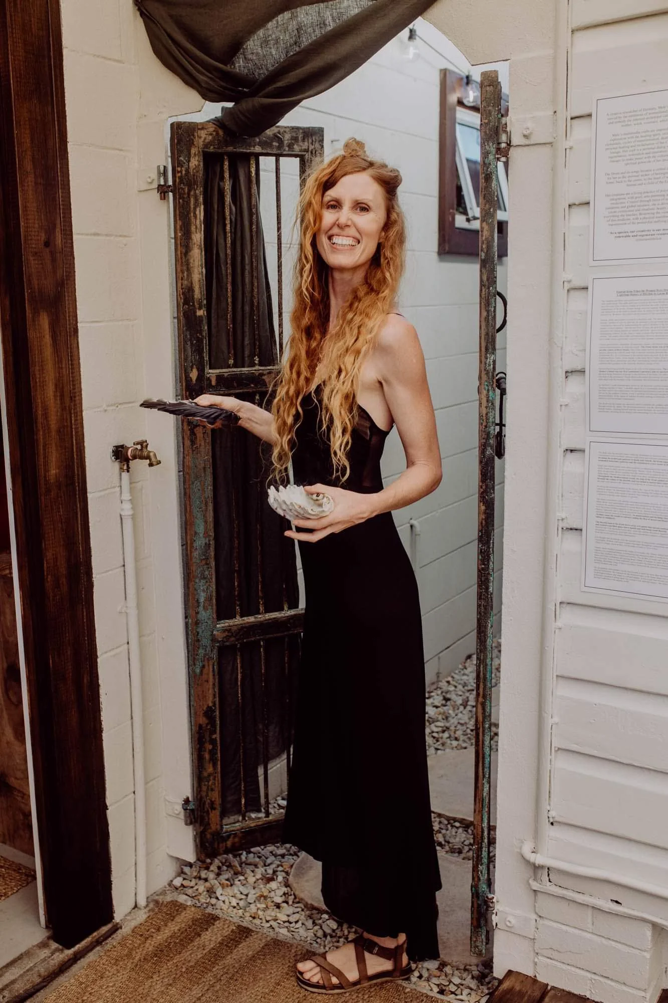 A woman with long, curly red hair wearing a black dress and sandals, standing in a doorway, smiling and holding a plate and a shell.